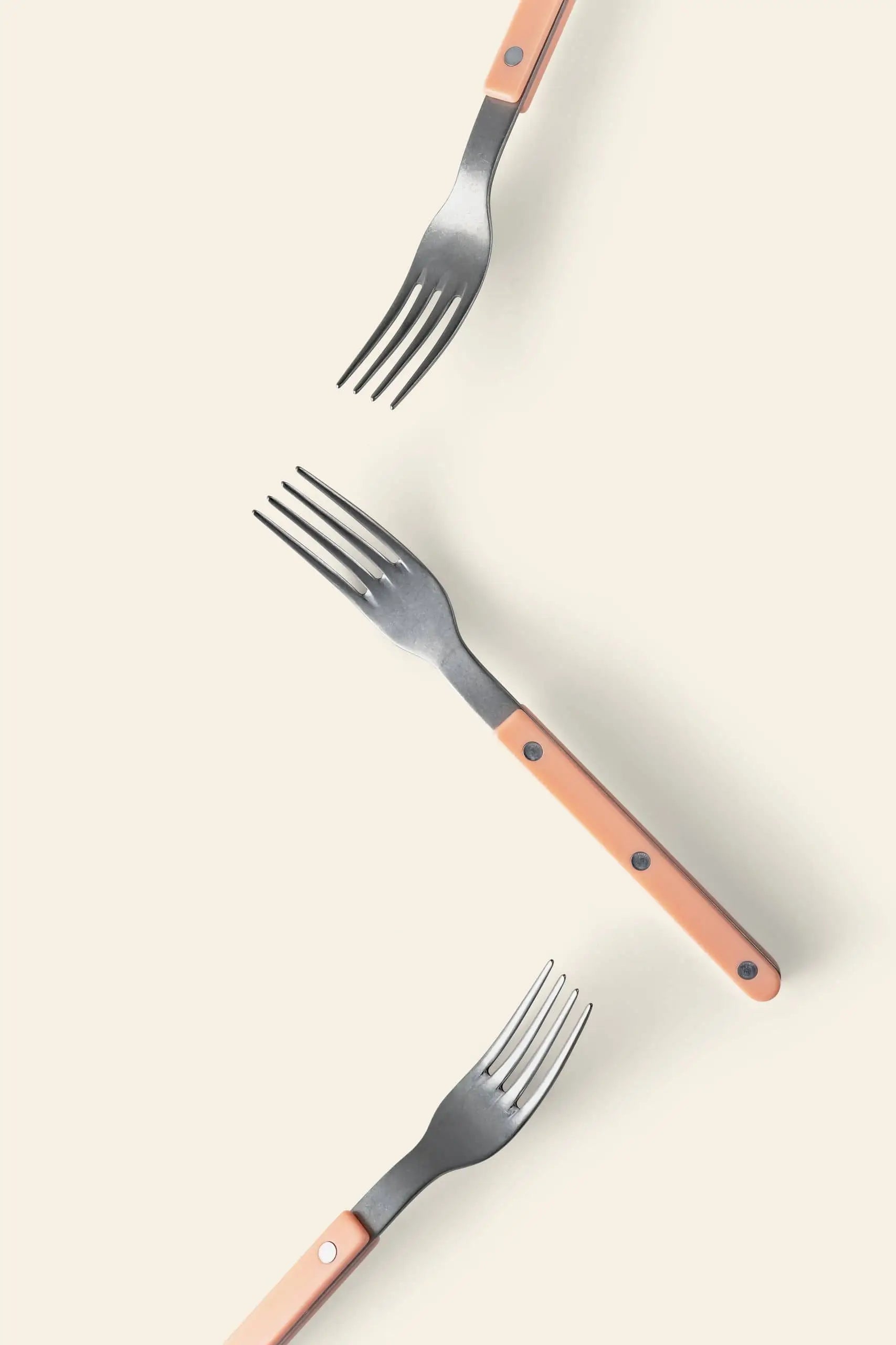 Three Sabre Bistrot Vintage Dinner Forks by Sabre, featuring peach-colored handles and stainless steel, are arranged diagonally on a light beige background, combining vintage charm with minimalist style for a striking visual.