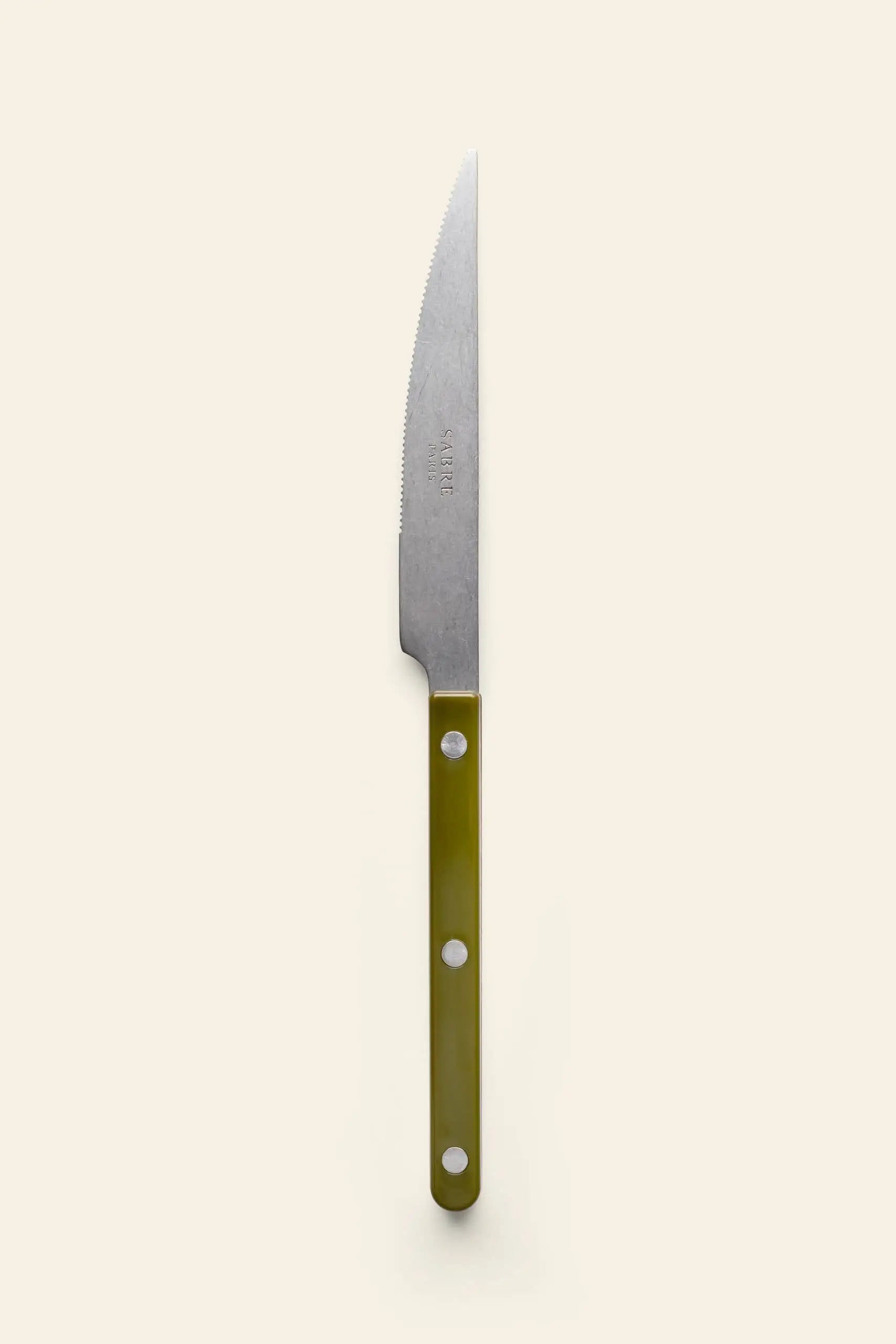 The Sabre Bistrot Vintage Dinner Knife by Sabre features a silver serrated blade and an olive green handle with three rivets, reflecting French bistrot style, shown on a plain light beige background.
