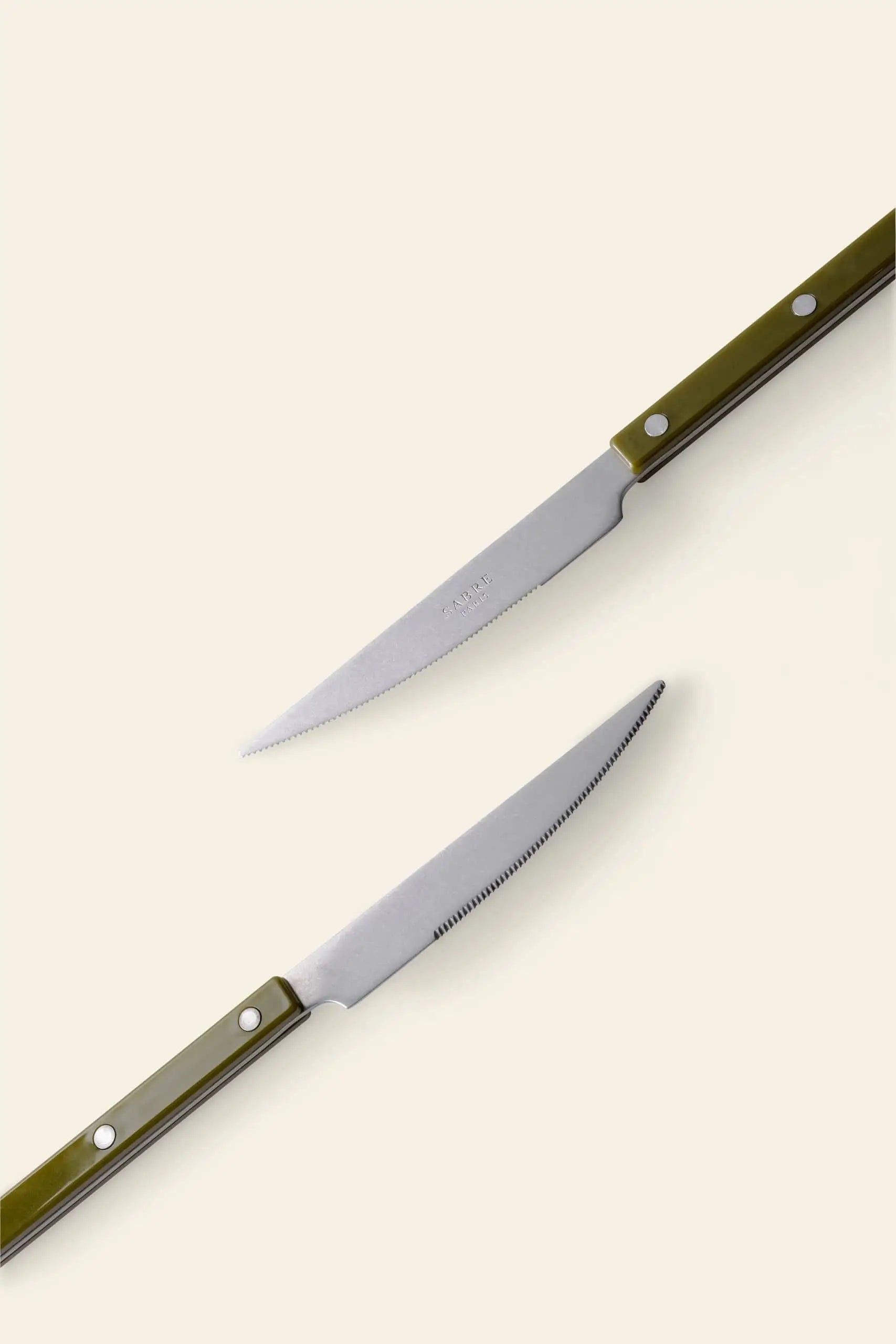 Two Sabre Bistrot Vintage Dinner Knives by Sabre, with olive green handles and silver rivets, are placed diagonally on a light beige background, their blades facing each other in classic French bistrot style.