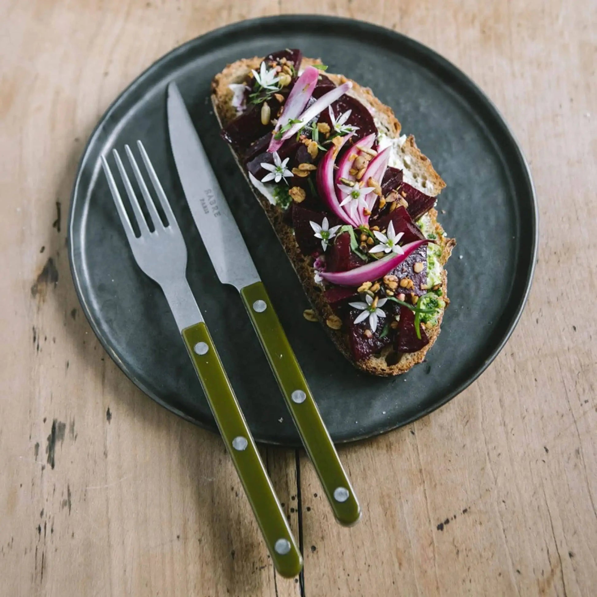A slice of bread with beetroot, red onion, herbs, edible flowers, and seeds sits on a dark plate beside a Sabre Bistrot Vintage Dinner Knife by Sabre on a wooden table.