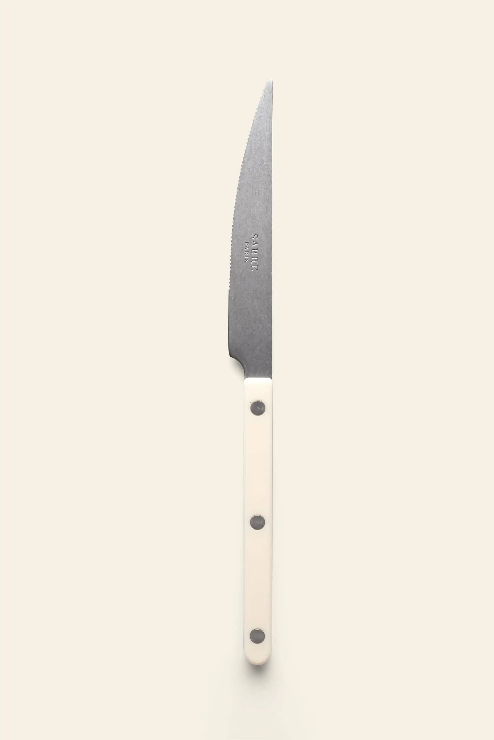 The Sabre Bistrot Vintage Dinner Knife by Sabre features a stainless steel serrated blade and a white handle with three metal rivets, displayed vertically on a light background—channeling classic French bistrot style.