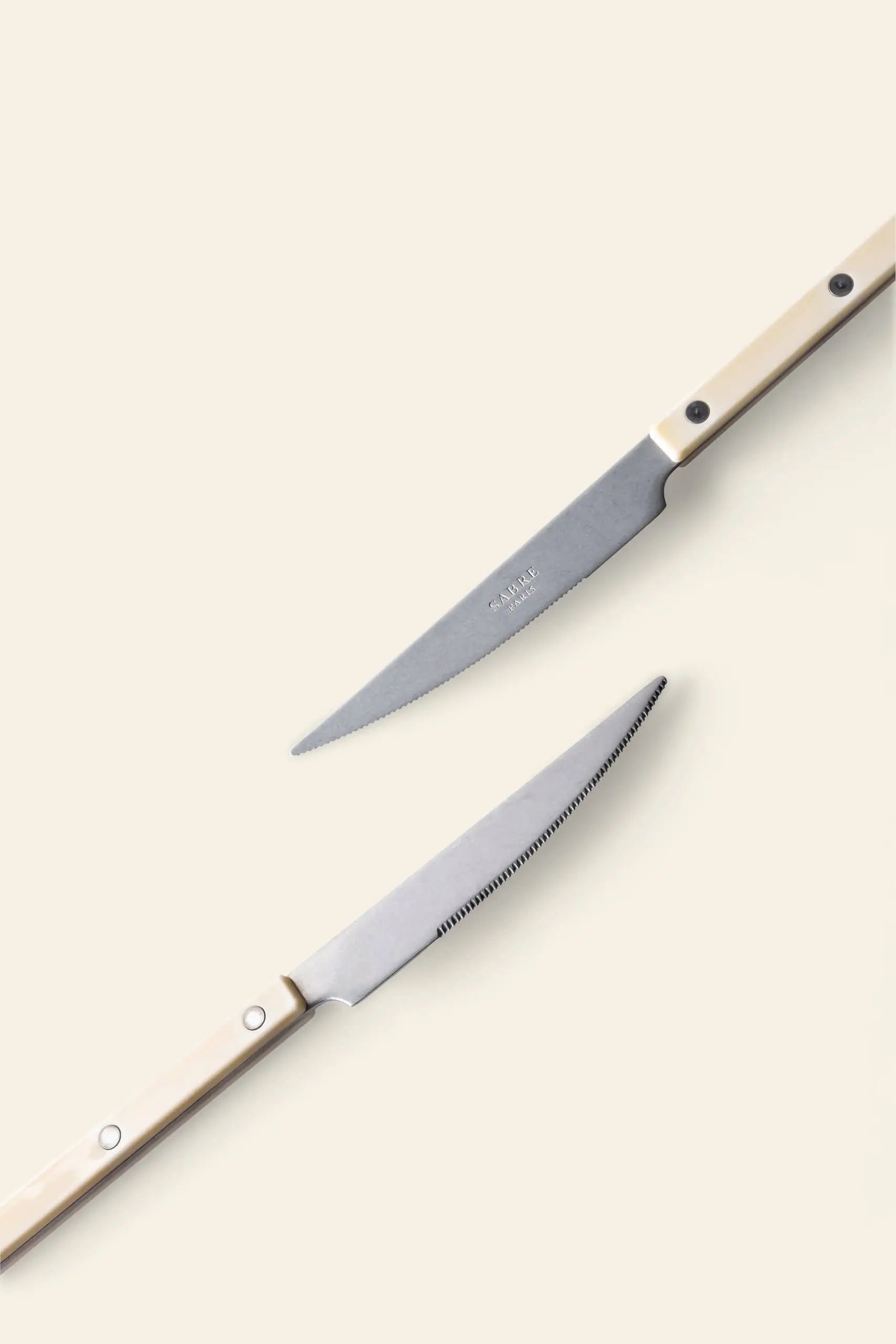 Two Sabre Bistrot Vintage Dinner Knives by Sabre, featuring light-colored handles and French bistrot-inspired design, are placed diagonally on a neutral background with blades pointing in opposite directions.