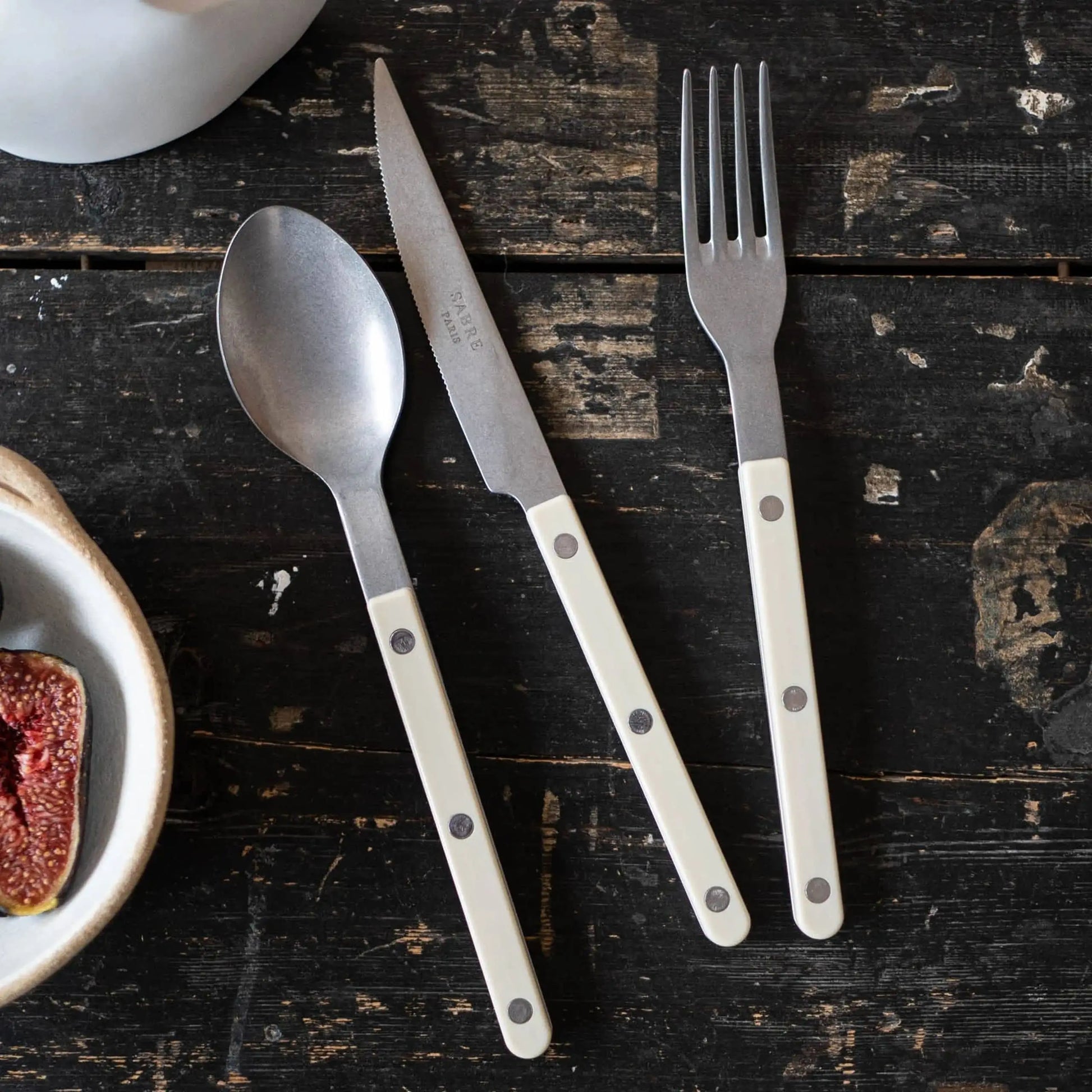 A spoon, a French bistrot style fork, and the Sabre Bistrot Vintage Dinner Knife by Sabre with white handles and metal rivets are neatly arranged on a dark wooden table. A bowl with a sliced fig is partially visible at the left edge.