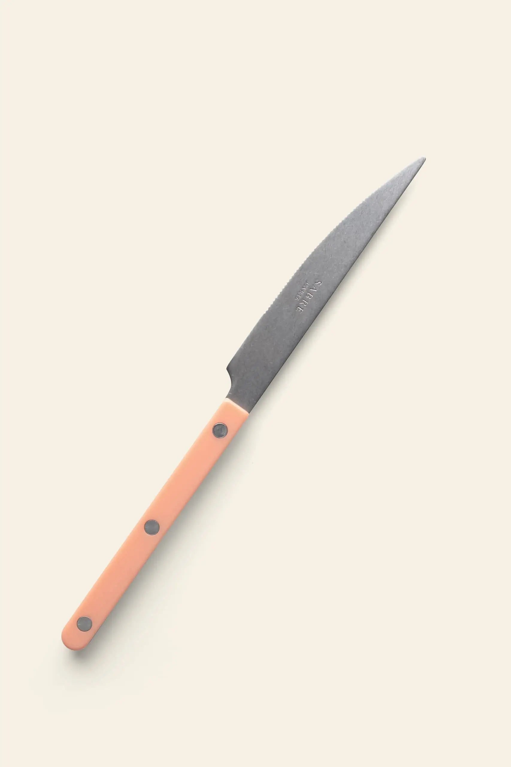 The Sabre Bistrot Vintage Dinner Knife by Sabre features a pointed dark blade and light pink handle with three metal rivets, capturing classic French bistrot style. Displayed against a plain, light beige background.