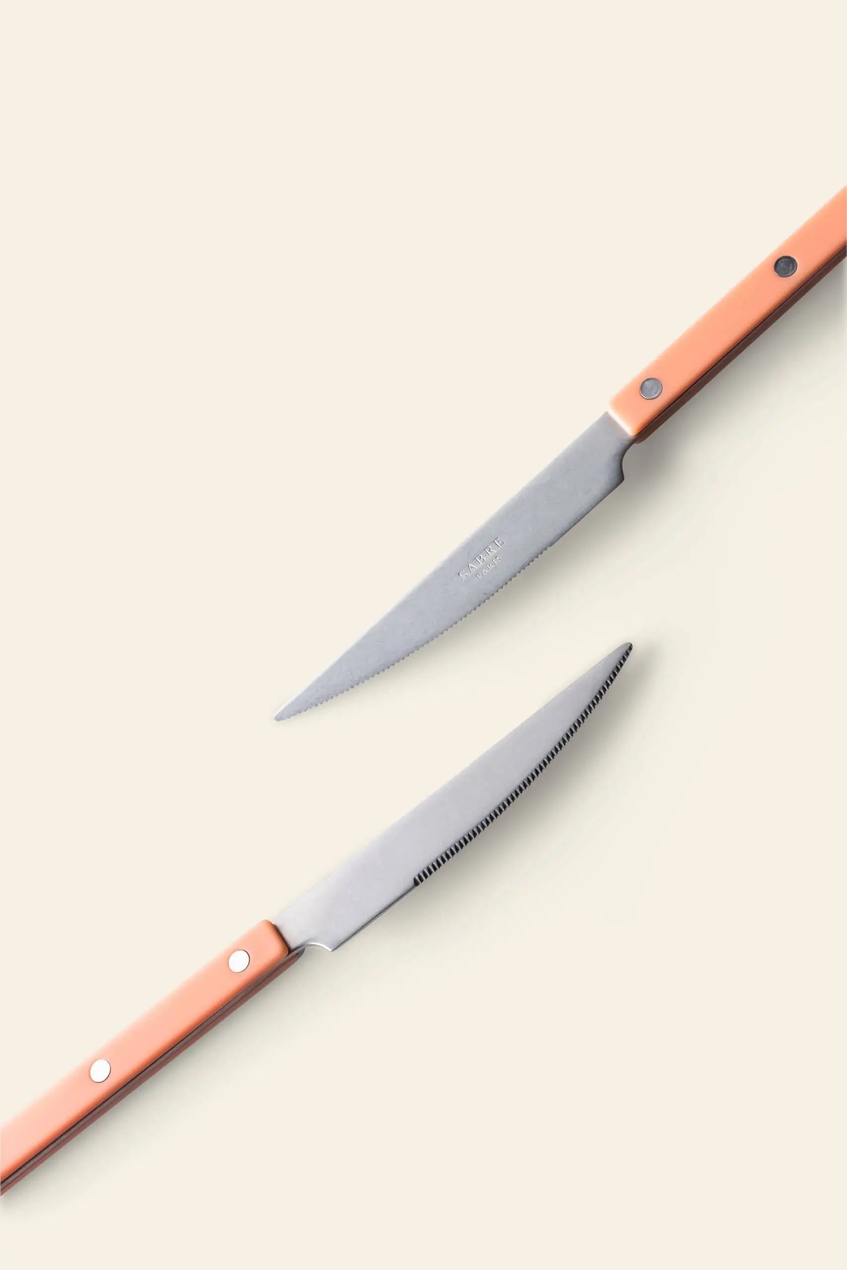 Two Sabre Bistrot Vintage Dinner Knives by Sabre, featuring silver blades and orange handles, are arranged diagonally on a light beige background for a French bistrot vibe. One rustic knife faces up, the other points down.