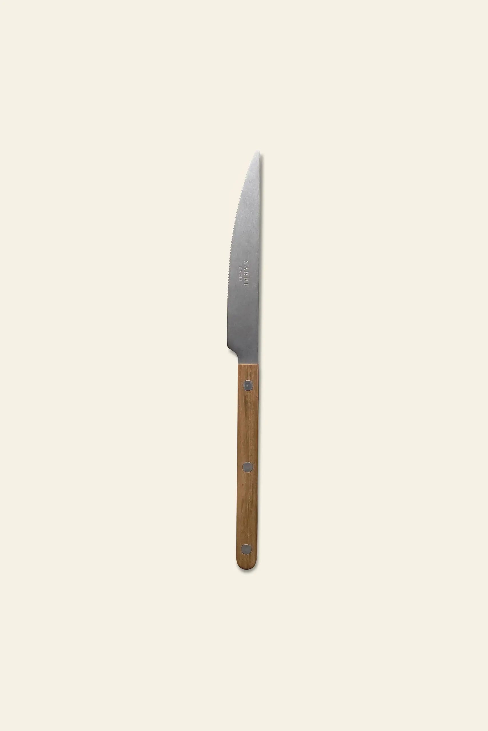 The Sabre Bistrot Vintage Dinner Knife by Sabre features a stainless steel serrated blade and a wooden handle, presented on a plain light beige background.