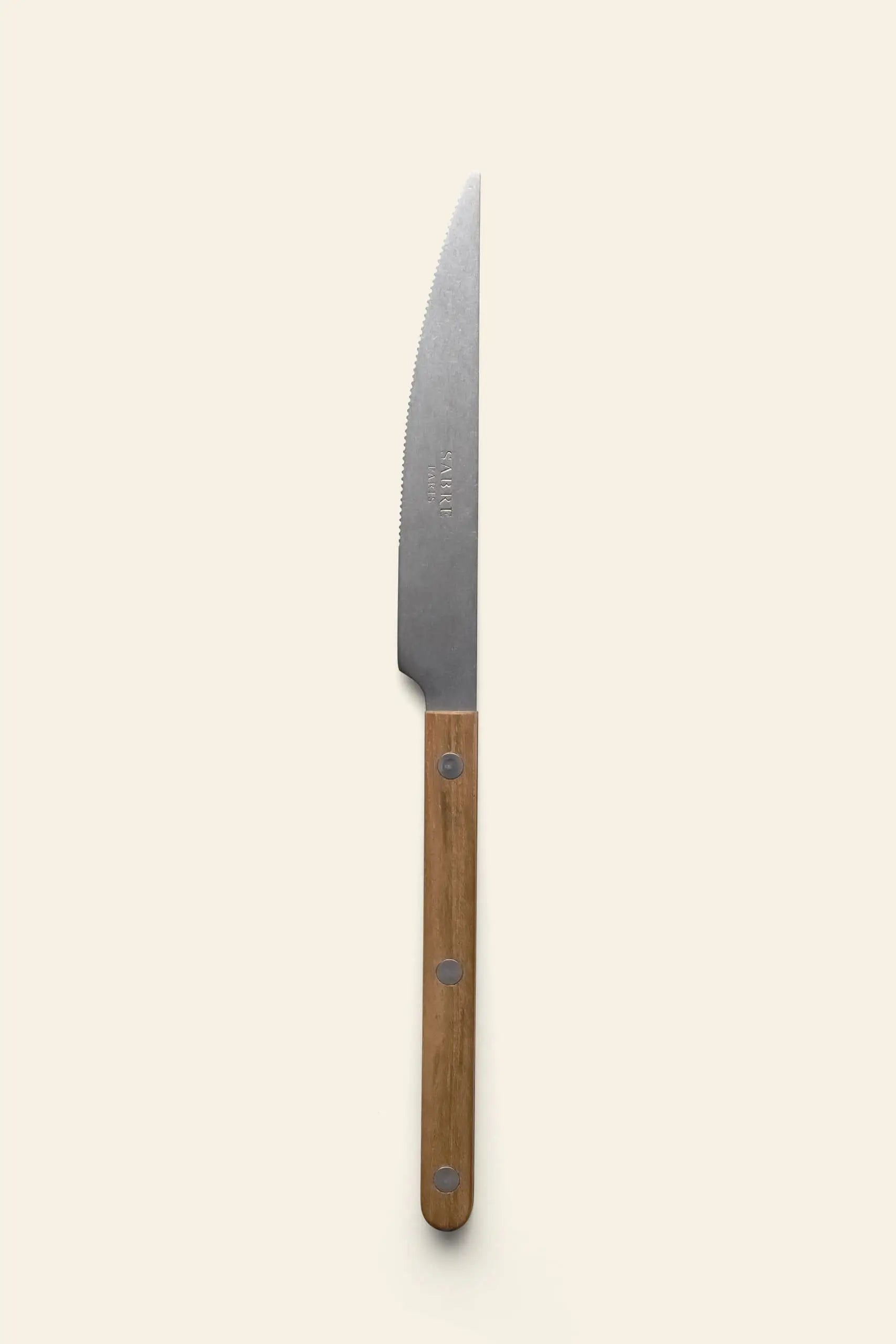 The Sabre Bistrot Vintage Dinner Knife by Sabre, featuring a serrated blade and wood handle with three metal rivets, stands vertically on a plain light beige background, reflecting classic French bistrot style.