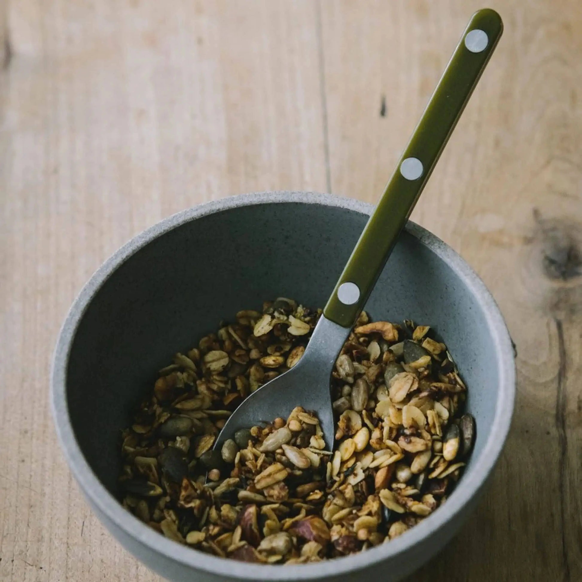 A grey bowl filled with granola sits on a wooden surface. Inside rests the Sabre Bistrot Vintage Soup Spoon by Sabre, featuring a green handle with white dots, evoking French café charm.