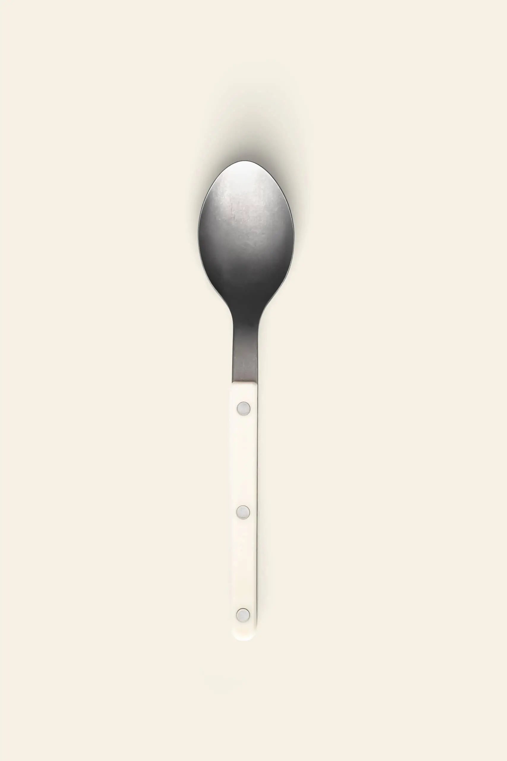 The Sabre Bistrot Vintage Soup Spoon by Sabre features a white handle with three silver rivets and classic stainless steel design, photographed on a light beige background to evoke the timeless charm of a French cafe.