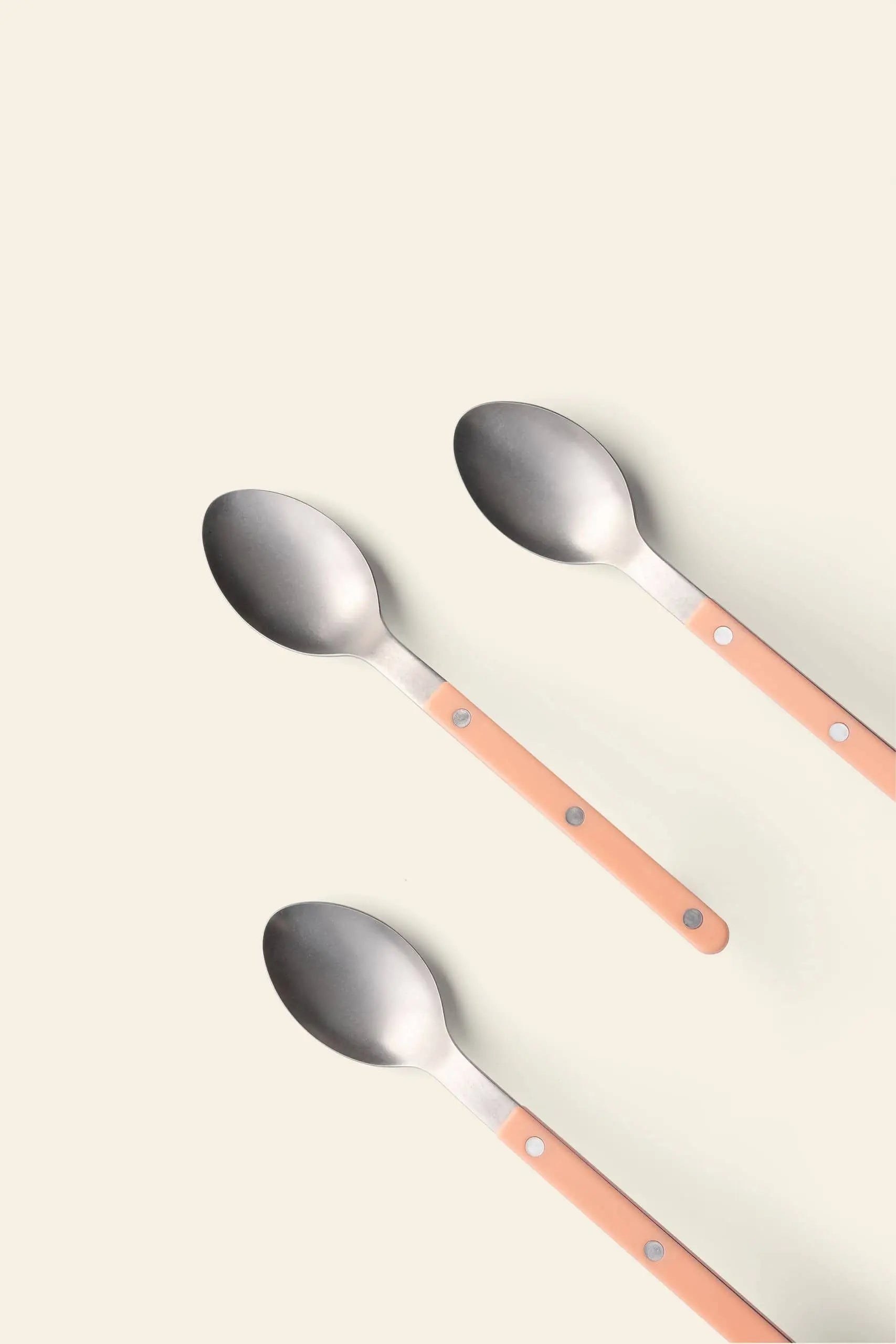 Three Sabre Bistrot Vintage Soup Spoons by Sabre, featuring stainless steel heads and light peach handles with metal rivets, are arranged diagonally on a plain beige background, evoking classic French café charm.