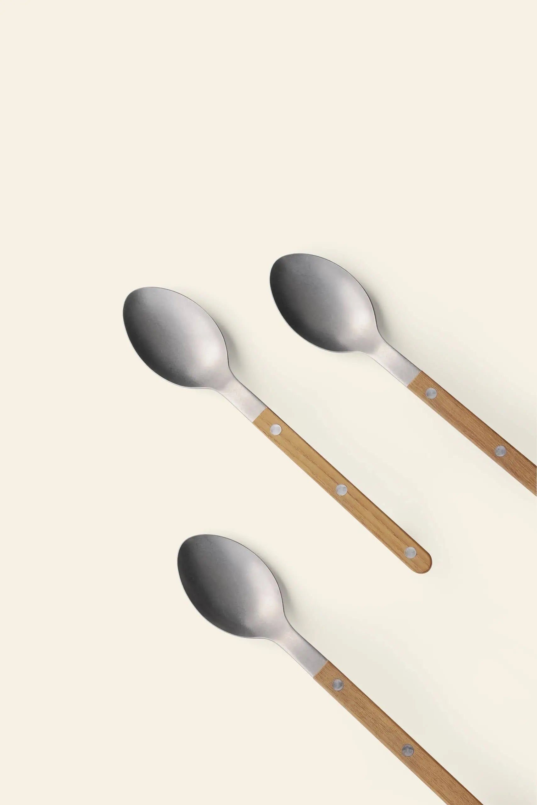 Three Sabre Bistrot Vintage Soup Spoons by Sabre, each with a stainless steel bowl and a wooden handle, are arranged diagonally on a light cream background—capturing the timeless charm of French café style.