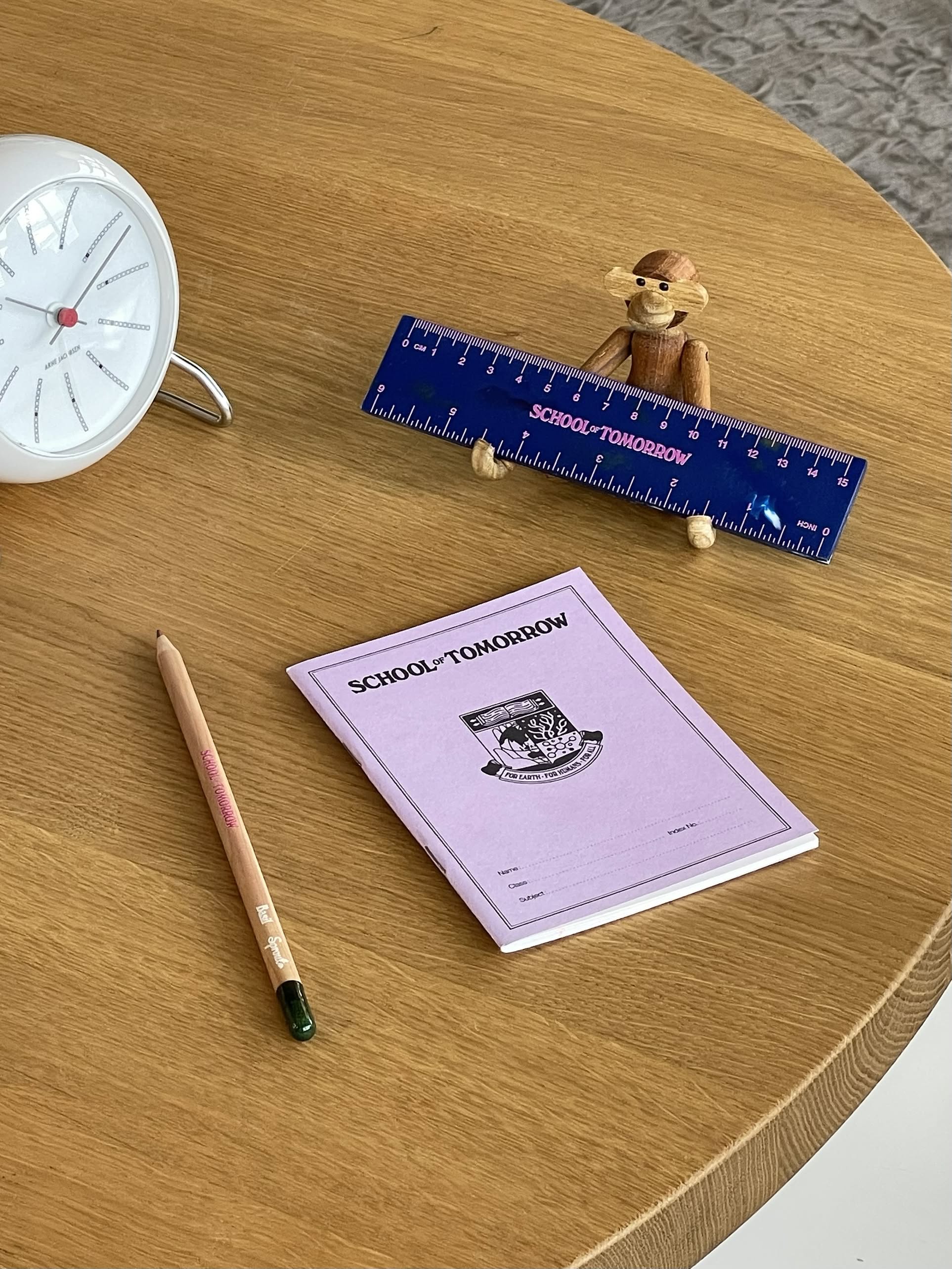 A wooden table holds a clock, a pencil, and the School of Tomorrow 15cm Ruler—crafted from recycled bottle caps—displayed by a small wooden monkey beside a purple SCHOOL TOMORROW booklet with its unique design.
