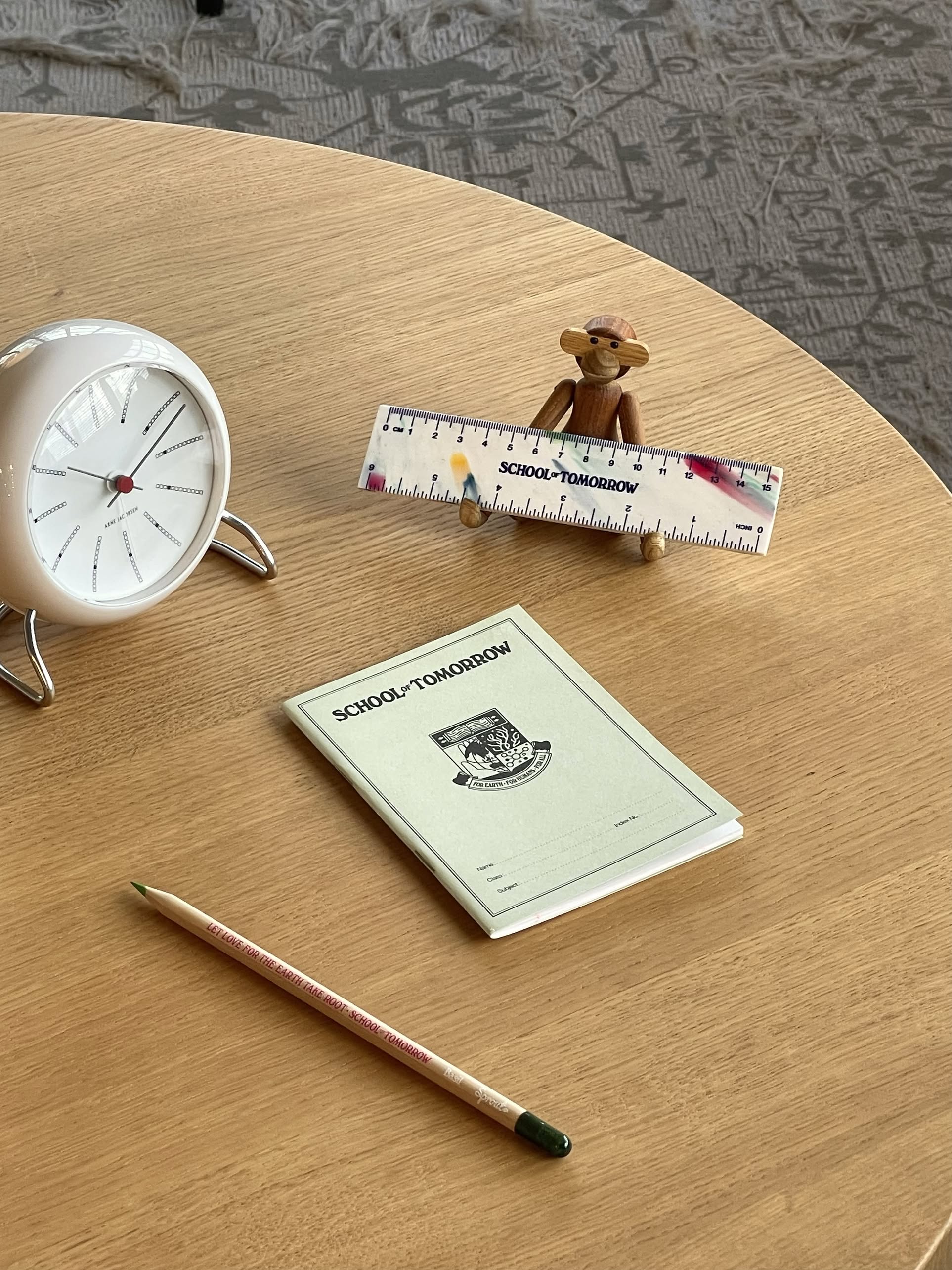 A round clock, pencil, booklet with School of Tomorrow design, small toy monkey, and a sustainably crafted School of Tomorrow 15cm Ruler are arranged on a wooden table with a patterned carpet in the background.