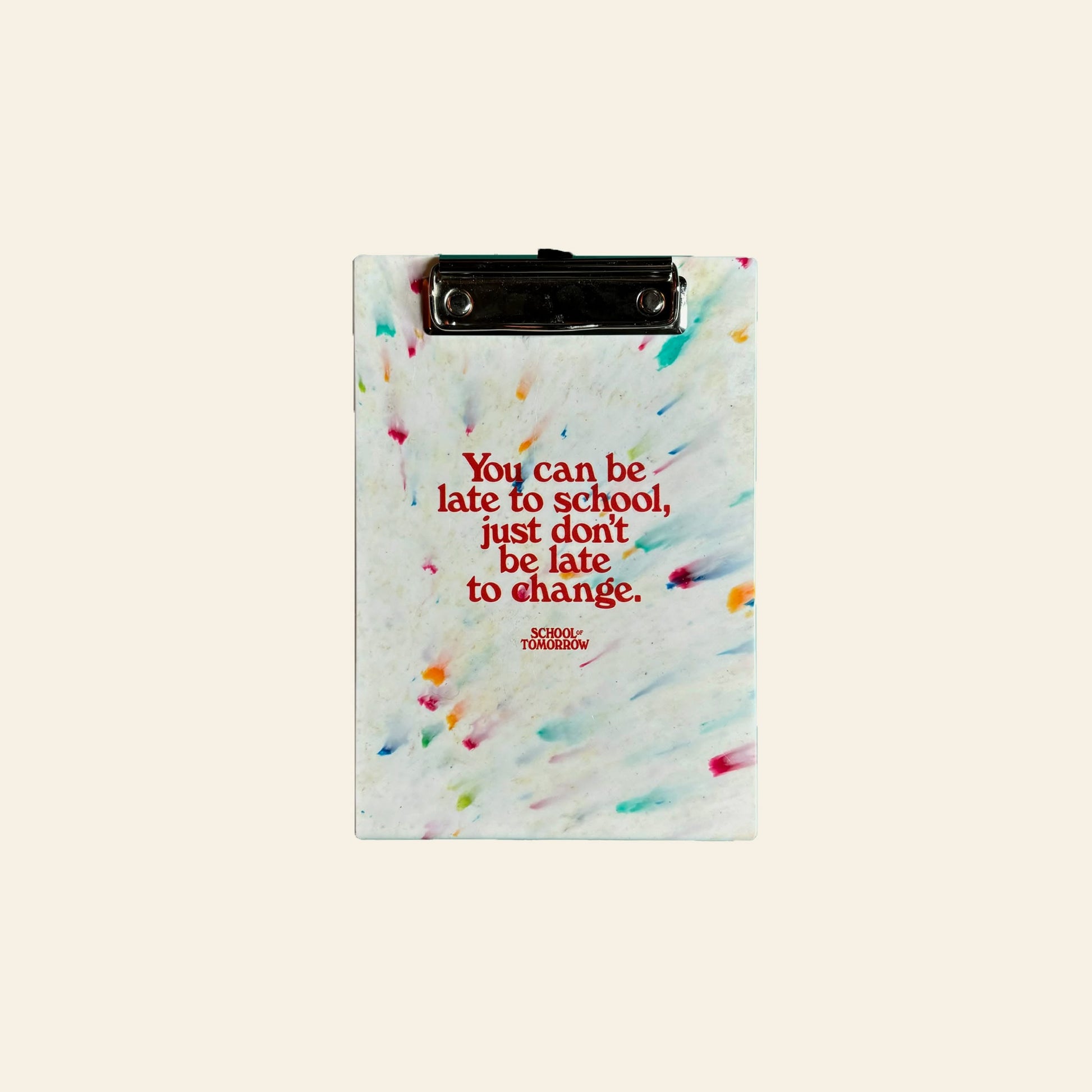The School of Tomorrow A5 Clipboard - White by School of Tomorrow is a small clipboard made from recycled bottle caps, featuring colorful speckles, a metal clip, and red text: You can be late to school, just don’t be late to change.