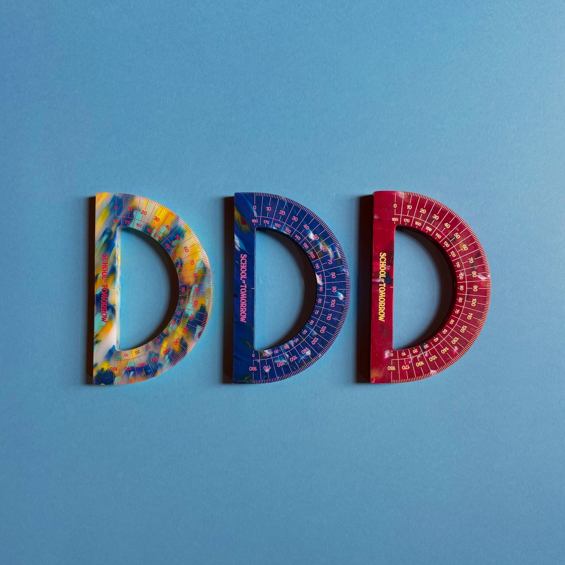 Three School of Tomorrow Math Instruments Protractors—yellow, blue, and red—are side by side on a blue background. Crafted from recycled HDPE plastic, these eco-friendly tools are D-shaped and from School of Tomorrow.