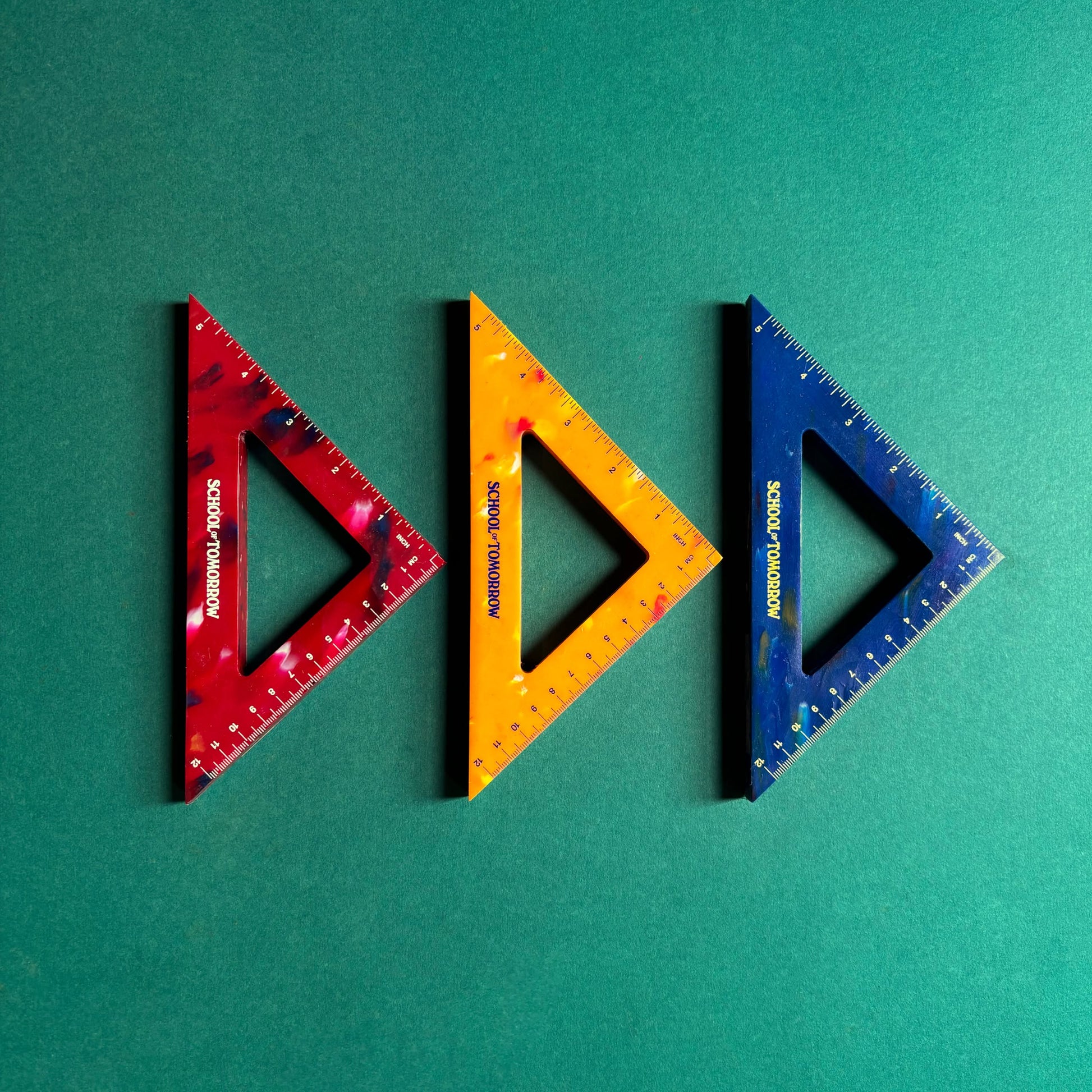 Three School of Tomorrow Math Instruments Triangles in red, yellow, and blue are arranged side by side on a green background. Made from recycled bottle caps, each features measurement markings and the School of Tomorrow brand name.