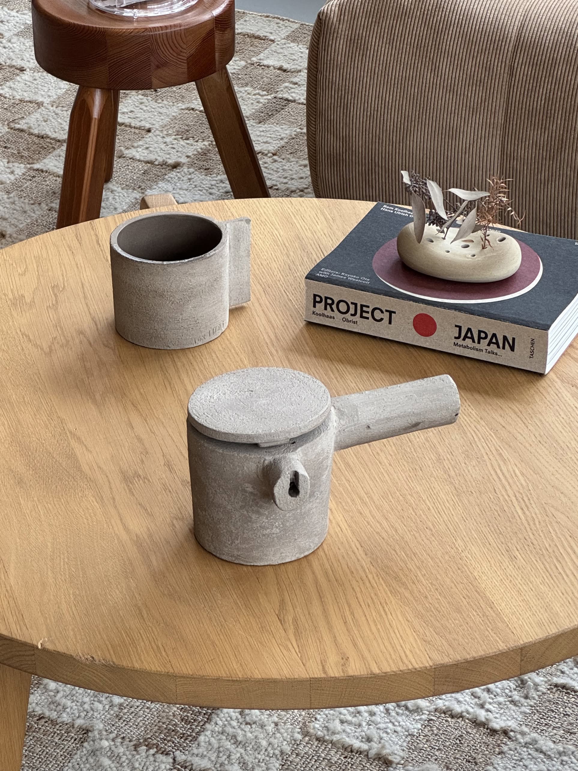 A round wooden table features a Serax Fck Teapot N°3 in grey brown, two Urbanistic Ceramics cups, and the book “Project Japan.” A decorative sculpture, brown stool, and patterned rug complete the modern concrete-finished space.
