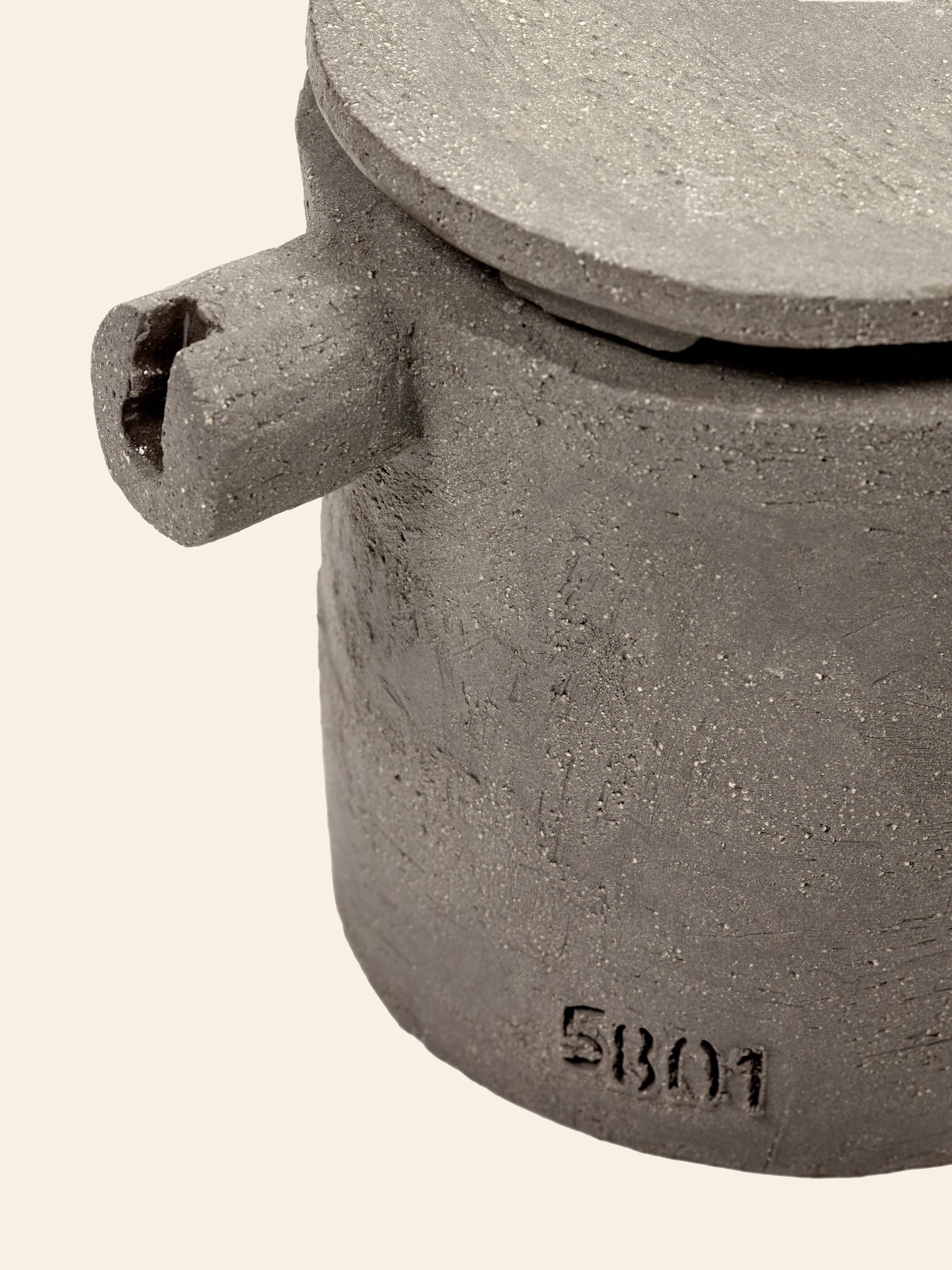 A close-up of the Serax Fck Teapot N°3 - Grey Brown by Serax shows its round, concrete form with lid and protruding handle, stamped “5801” near the base. The urban ceramics design stands out against a plain light background.
