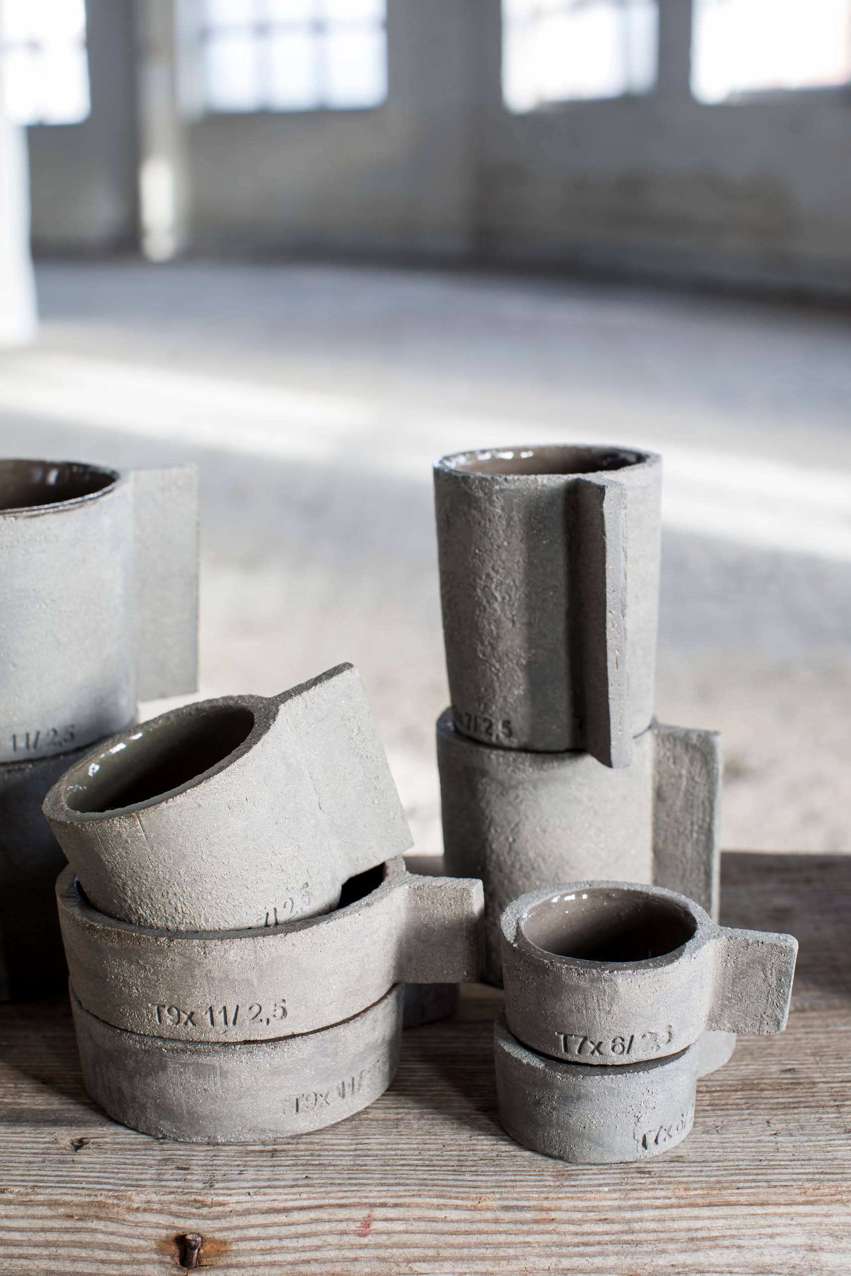 A collection of Serax Fck Teapot N°3 - Grey Brown ceramics with handles and stamped numbers is displayed on a wooden surface in a sunlit, industrial-style room with large windows.