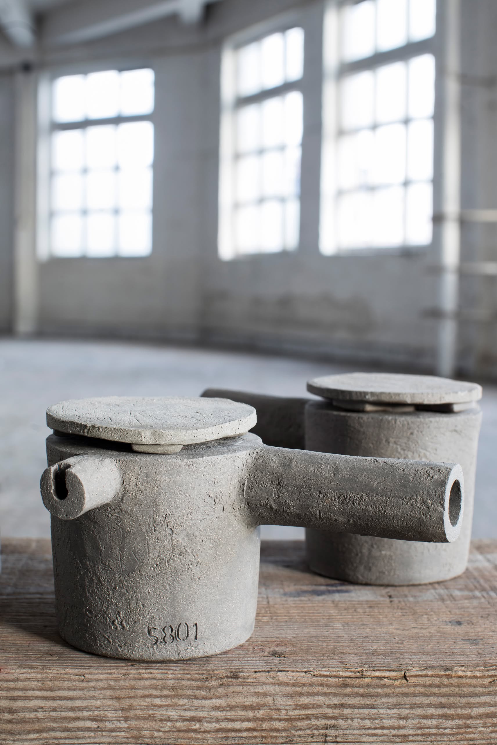 Two grey-brown Serax Fck Teapot N°3 by Serax with side spouts and flat lids are placed on a wooden surface in a spacious, bright room with tall windows and a concrete floor.