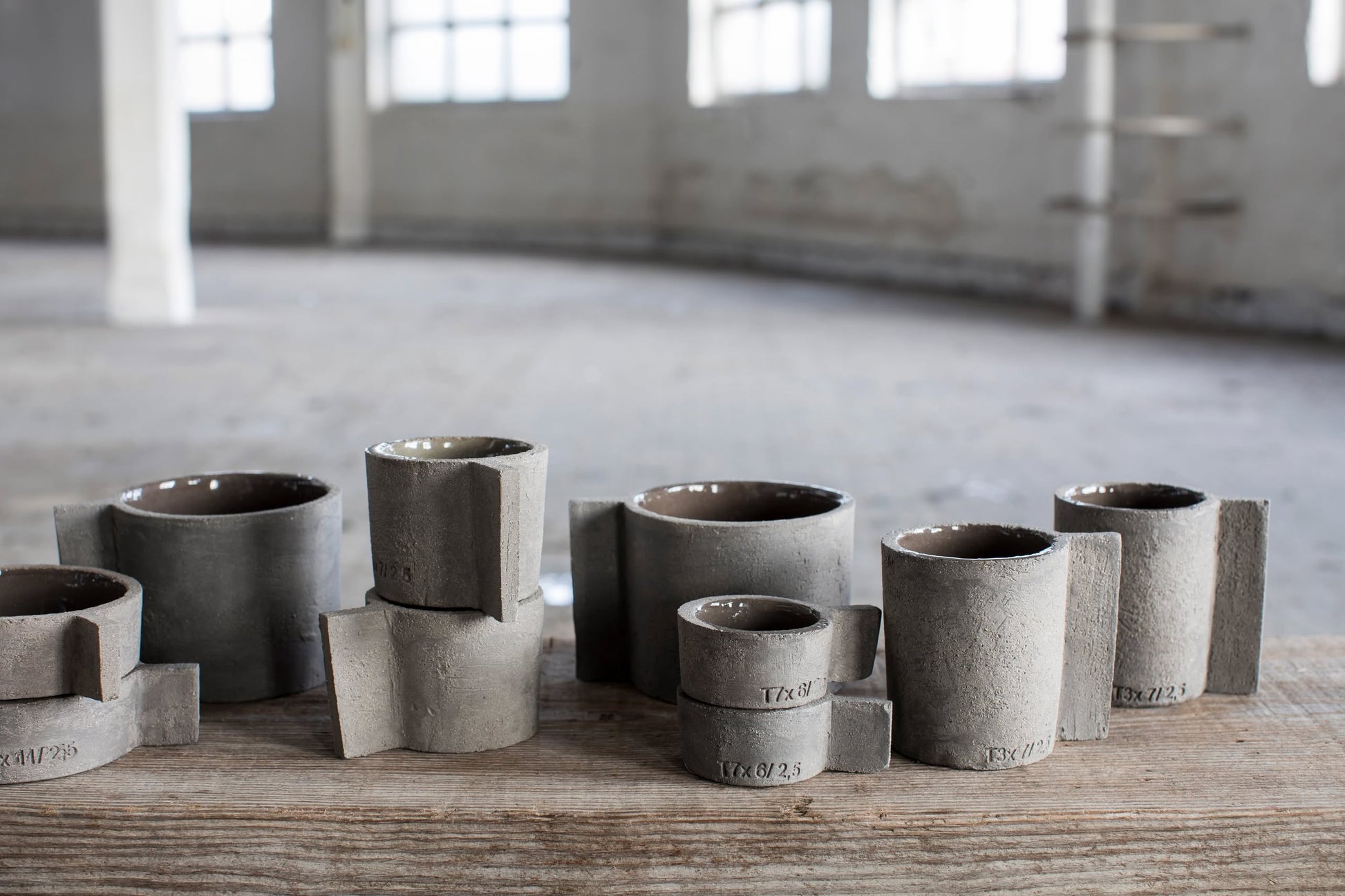 The Serax Fck Teapot N°3 in grey brown by Serax is displayed on a wooden surface in a bright, industrial-style room with large windows, highlighting its handmade, geometric design and unique concrete finish.