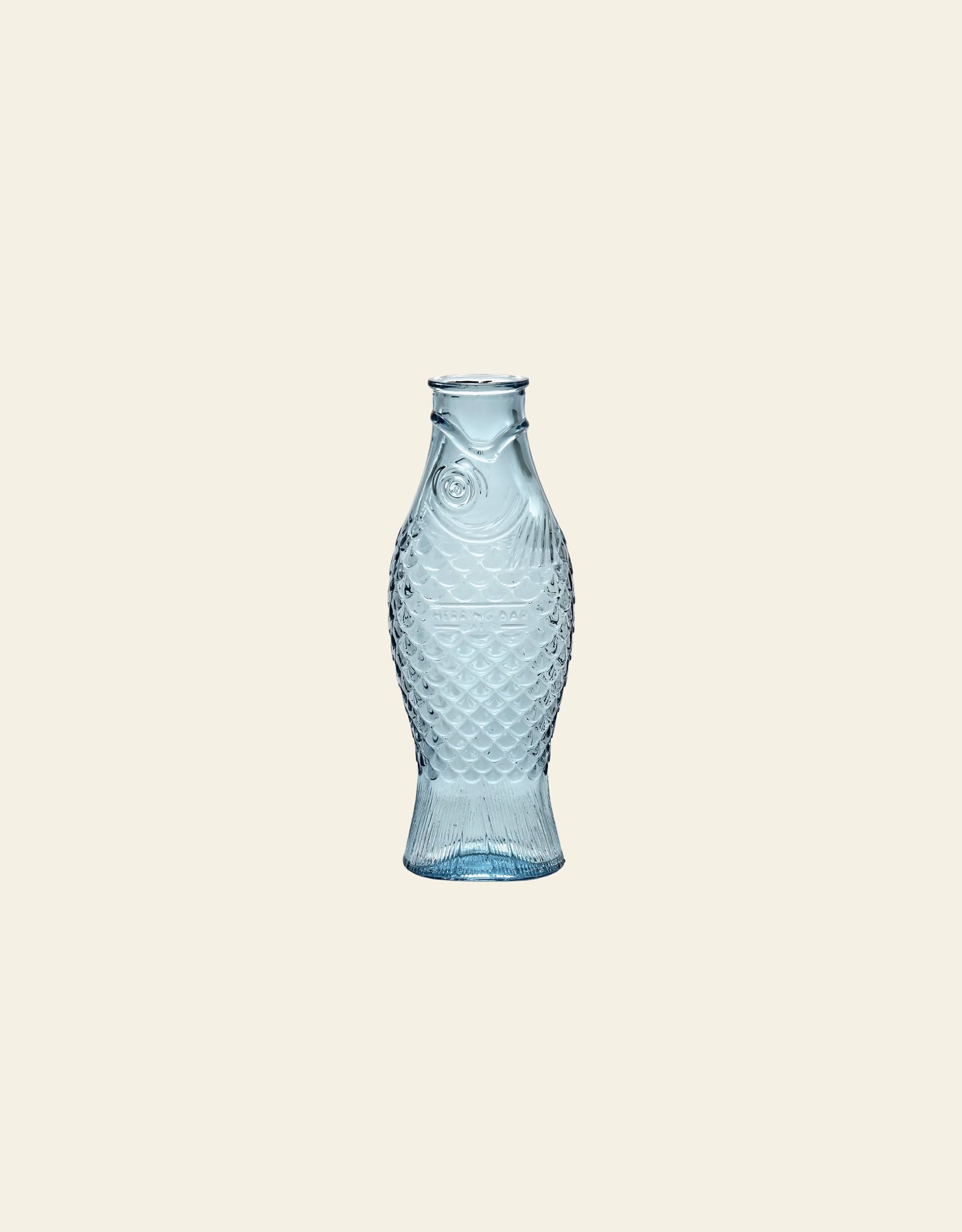 The Serax Fish & Fish Carafe by Serax, featuring textured scales and fins in transparent light blue glass, stands out as an eye-catching tableware piece against a plain beige background.