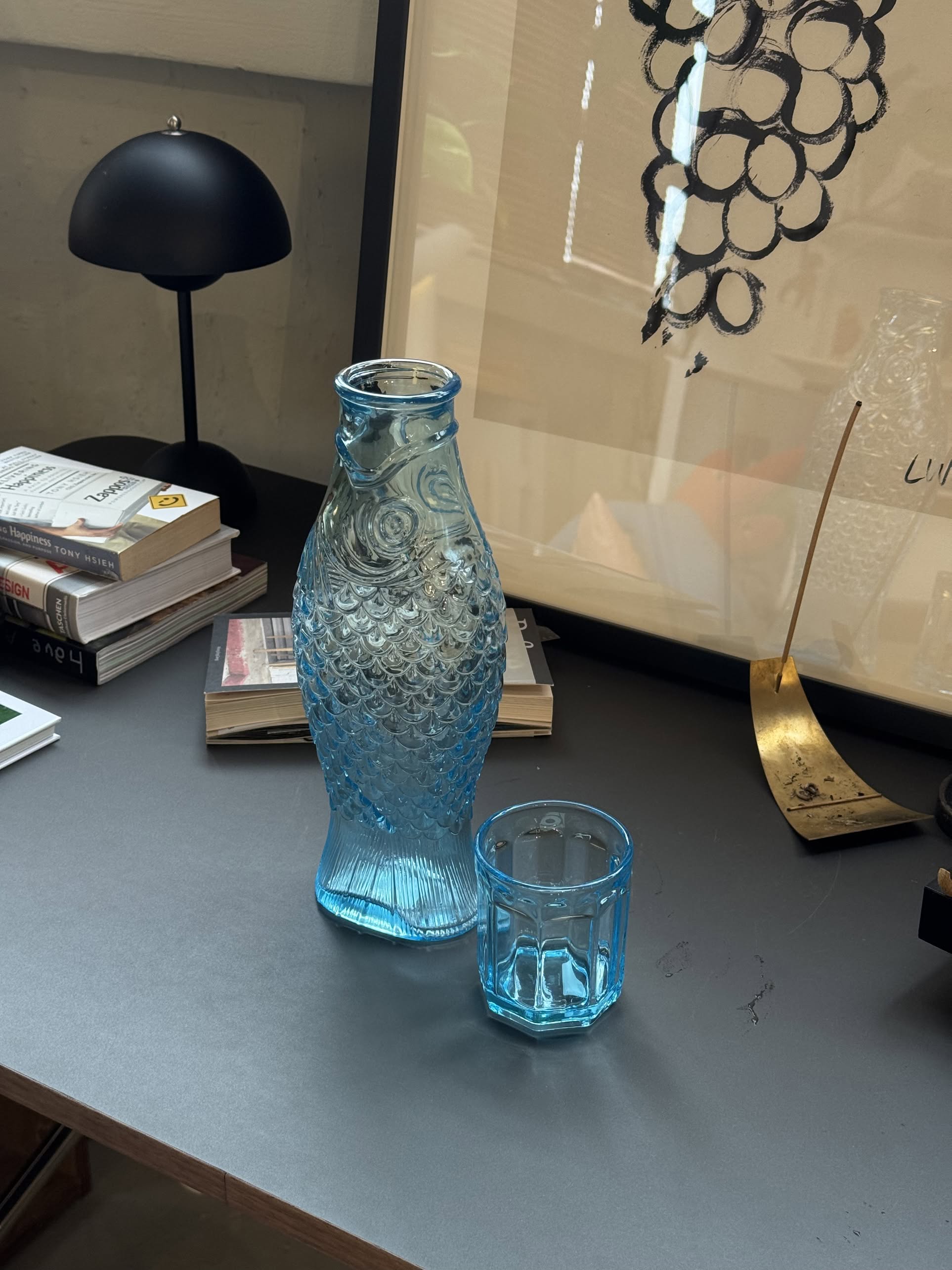 A blue Serax Fish & Fish Carafe by Serax sits beside a matching blue glass on a desk with books, a black lamp, and framed art—unique tableware to enhance your space.