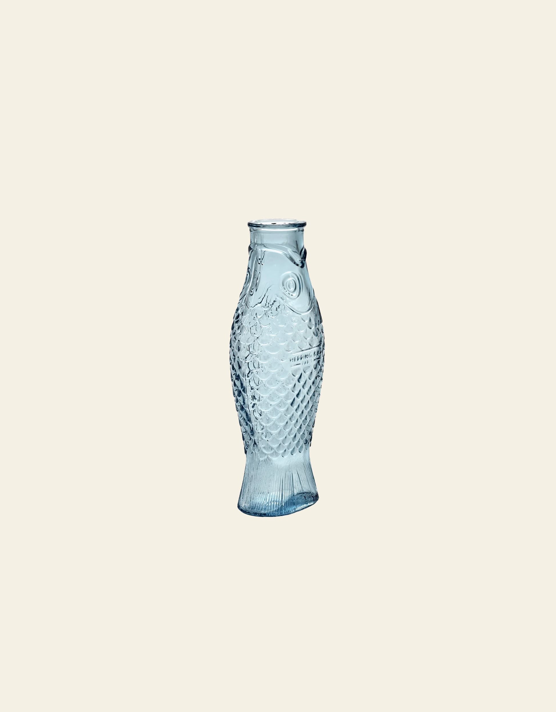 The Serax Fish & Fish Carafe by Serax, with its blue-tinted glass, detailed scales, and fins, stands out as a unique piece of tableware set against a plain light beige background.