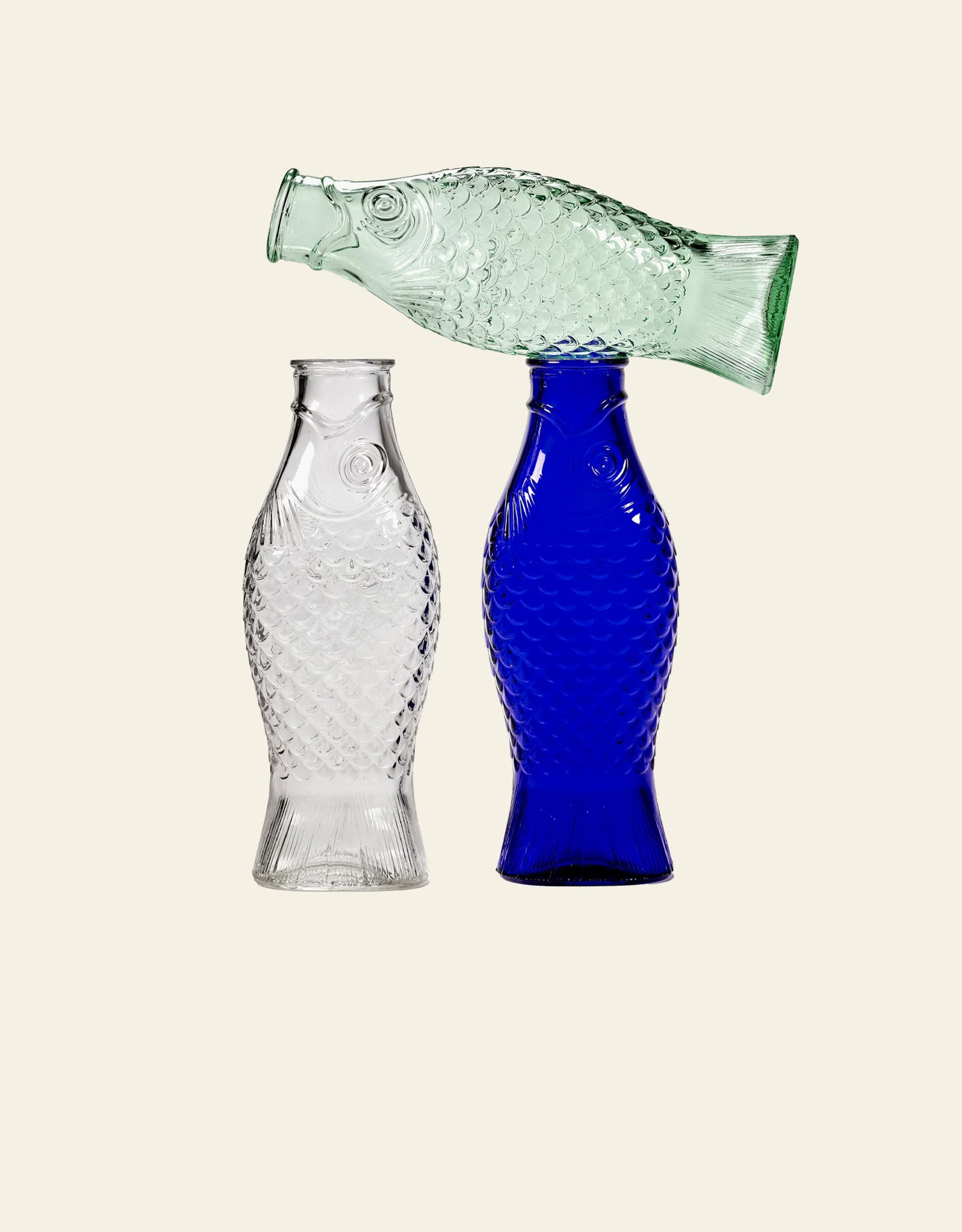 The Serax Fish & Fish Carafe set by Serax features three glass carafes—clear, blue, and light green—shaped like fish. The clear and blue stand upright, while the green rests horizontally on top for a unique table display.