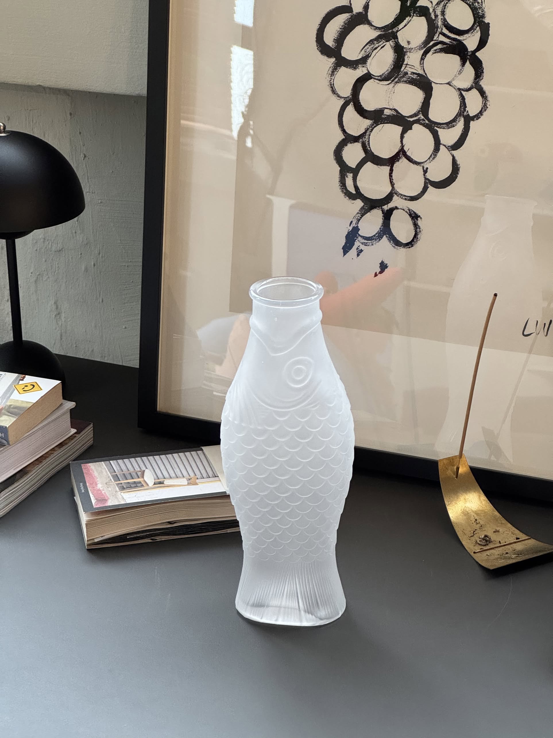 A frosted glass Serax Fish & Fish Carafe by Serax, shaped like a fish, rests on a dark table beside stacked books, a black lamp, gold ornament, and art with a grape-like design—ideal for unique tableware enthusiasts.