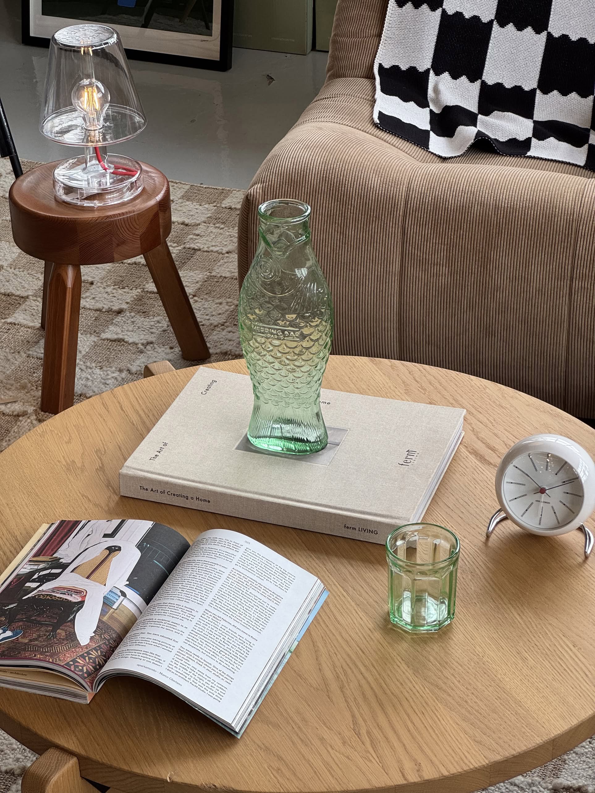 A wooden coffee table with a Serax Fish & Fish Carafe in green, tumbler, open magazine, large book, and white analog clock. In the background: striped blanket, brown sofa, stool, and lamp.