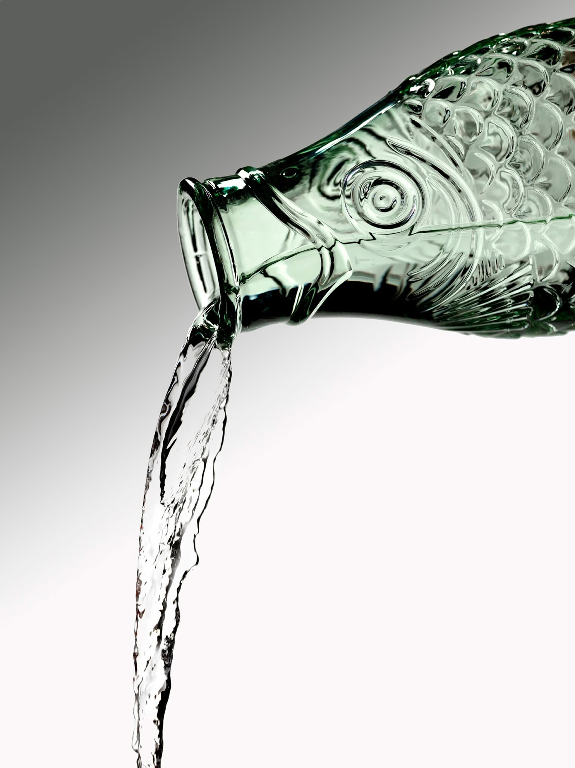 A green, fish-shaped glass Serax Fish & Fish Carafe by Serax is tilted, pouring water from its mouth against a gradient white and gray background.