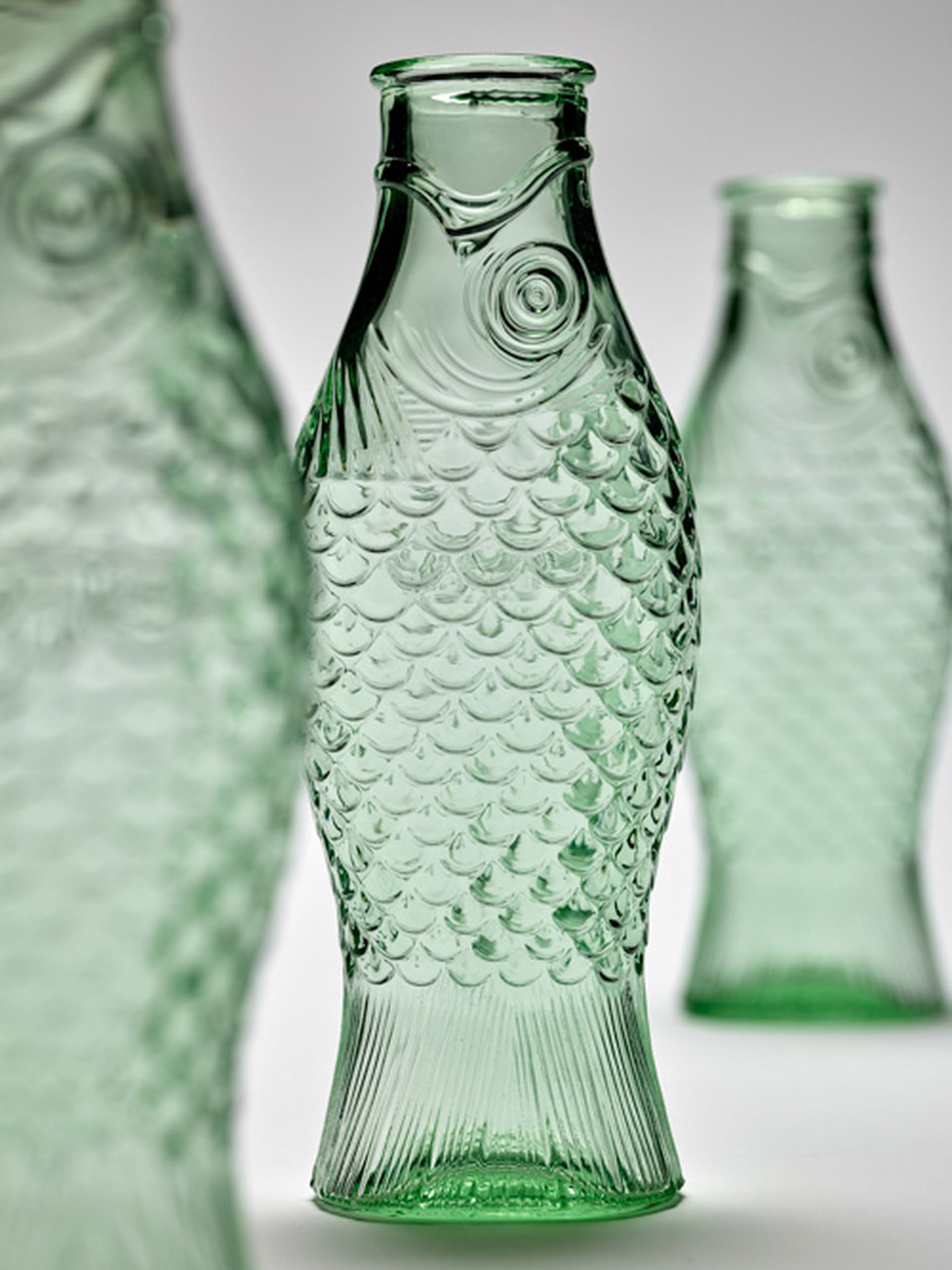 Three Serax Fish & Fish Carafes in green, shaped like textured fish with detailed scales and fins, stand on a plain light background. The central Serax bottle is in sharp focus while the others appear softly blurred behind.