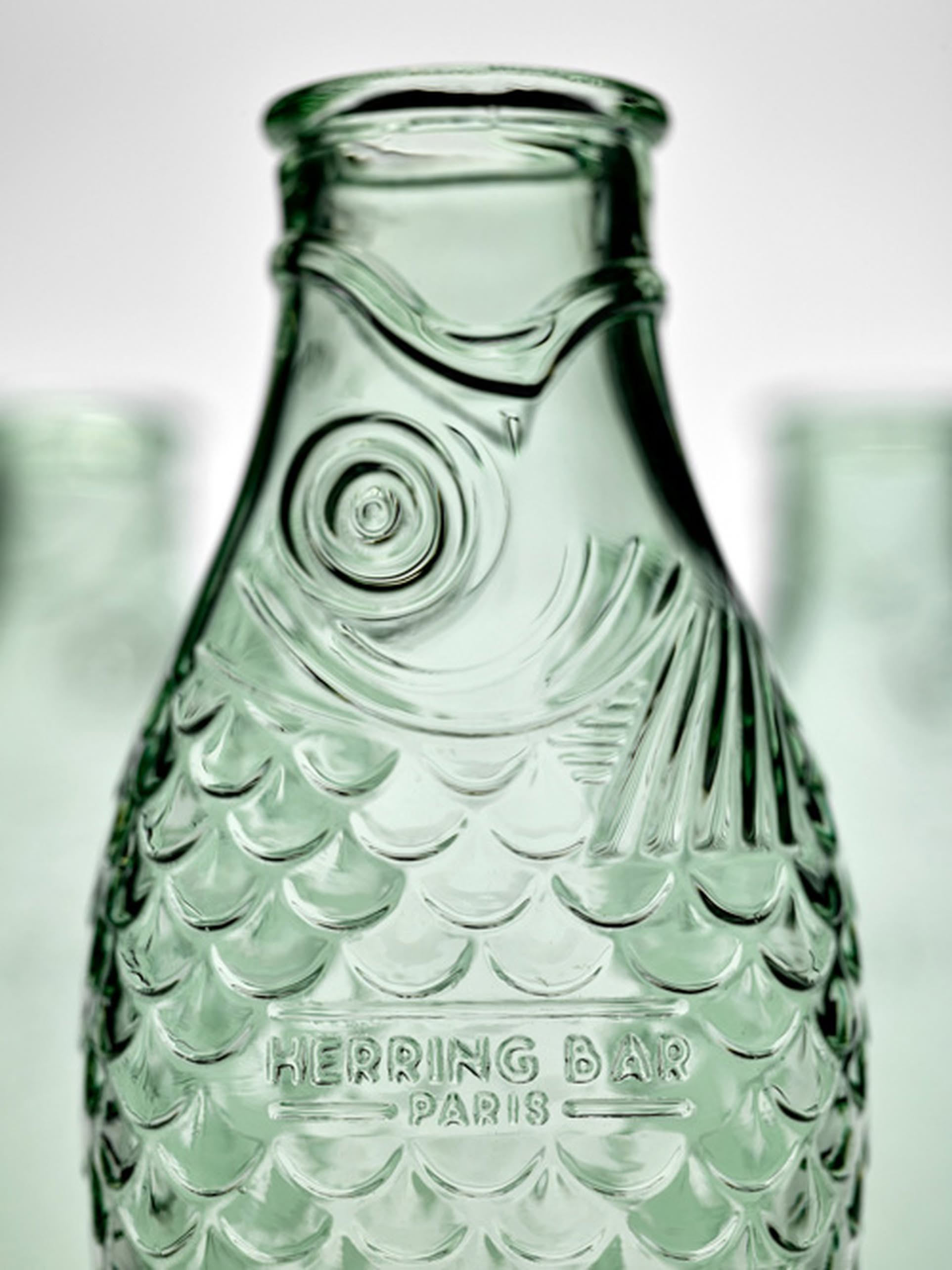 A green glass carafe from Serax’s Fish & Fish collection, shaped like a fish with embossed scales and HERRING BAR PARIS on its body. Designed by Paola Navone for Serax. Similar bottles appear softly in the background.