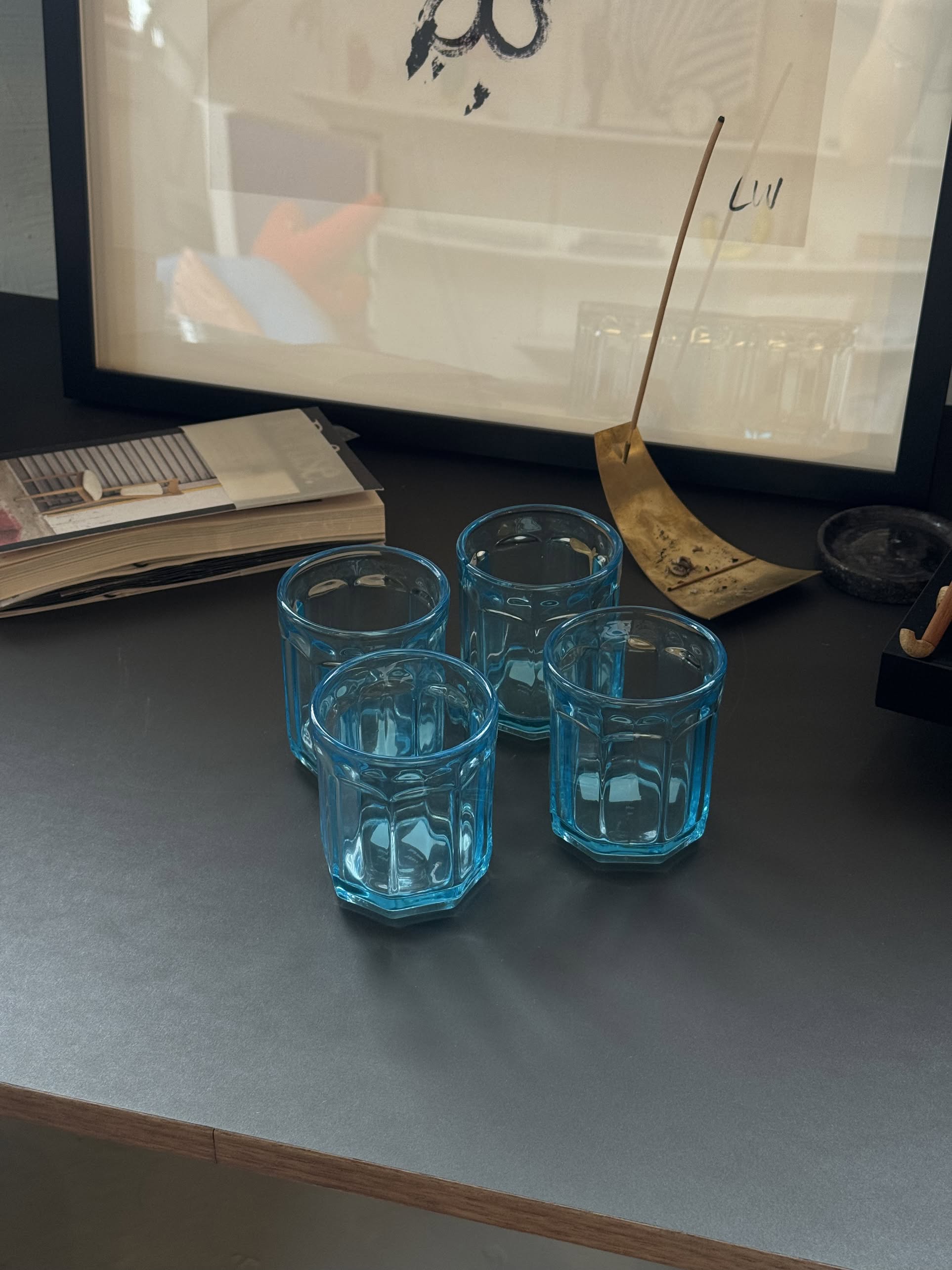 Four blue glass tumblers are arranged on a dark surface near a book, a brass incense holder, framed artwork, and a green Serax Fish & Fish Glass M carafe. Soft lighting highlights the display.