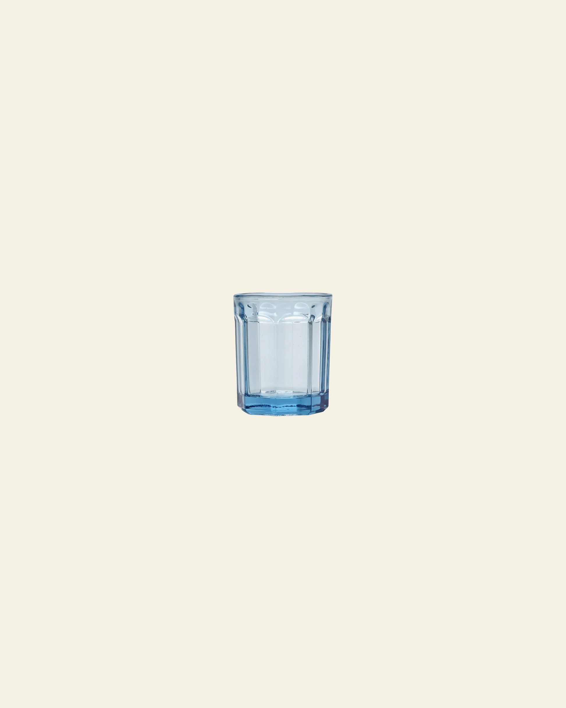 A blue-tinted glass tumbler, half-filled with water, is centered on a plain light beige background next to a green Serax Fish & Fish Glass M carafe by Serax.