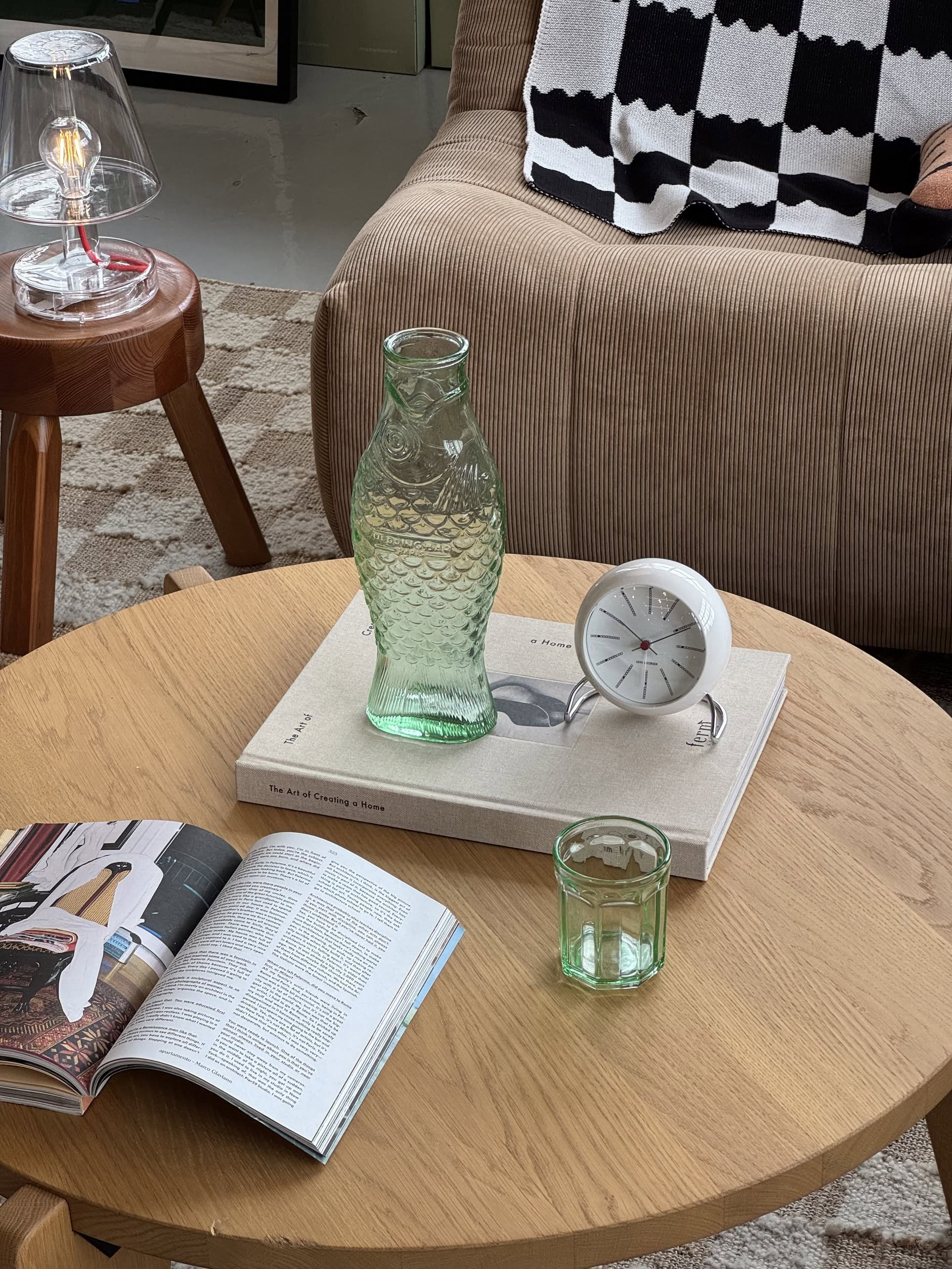 A wooden coffee table featuring the Serax Fish & Fish Glass M in green by Serax, a small clear glass, a tilted white clock, an art book, and an open magazine in a cozy living room setting.