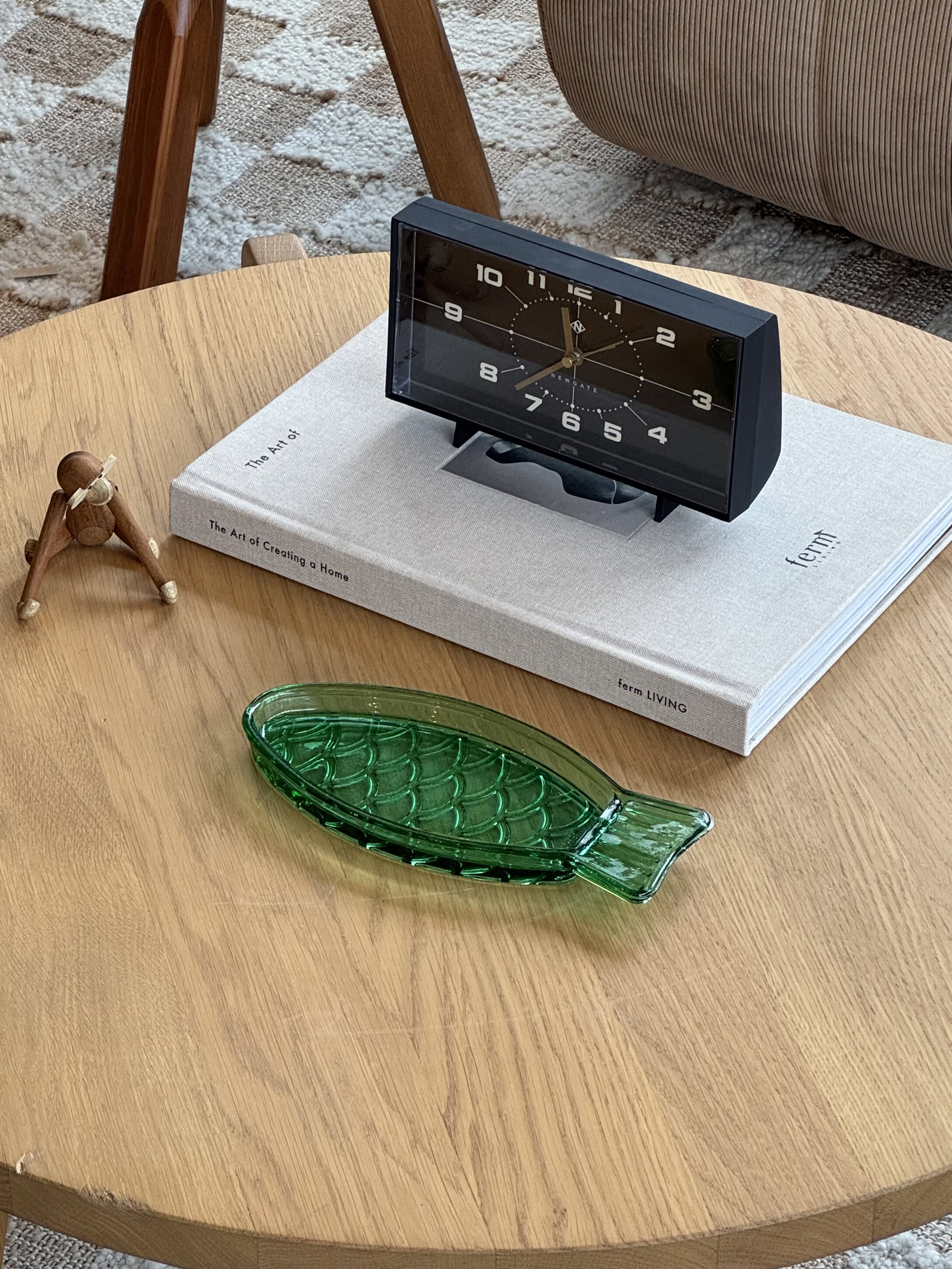 A round wooden table holds the Serax Fish & Fish Serving Dish N°1 - Green by Serax, a wooden figurine, the book The Art of Creating a Home, and a black vintage-style clock.