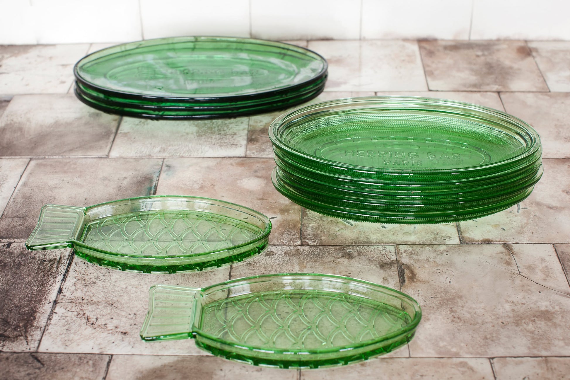 A set of vintage green glass plates, including round and oval stacks plus two Serax Fish & Fish Serving Dish N°1 - Green by Serax, arranged on a tiled surface.