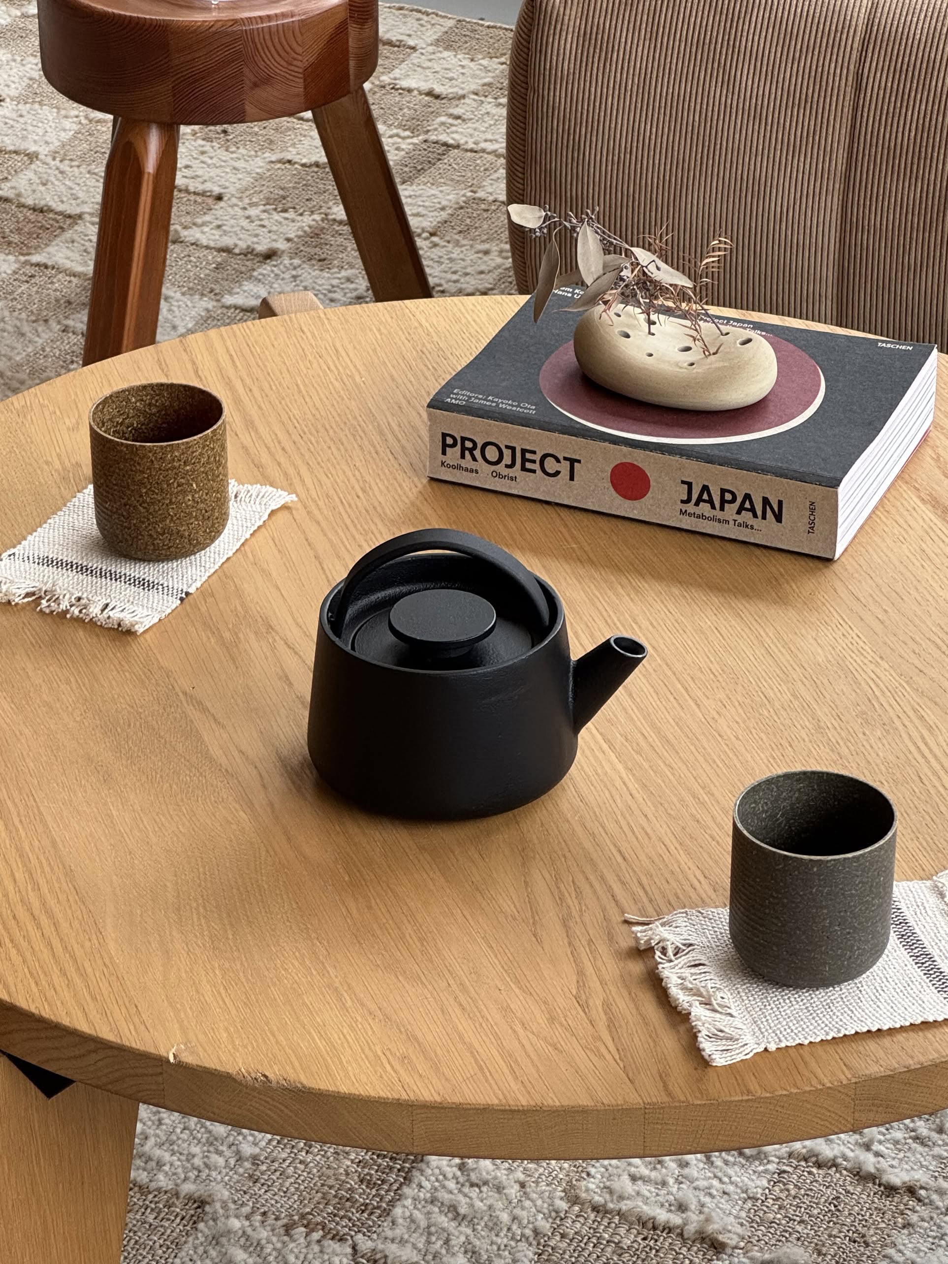 A round wooden table with two cups on coasters, a Serax Inku Cast Iron Teapot S - Black, and a book titled Project Japan. A small modern sculpture sits on the book, with a patterned rug beneath the table.