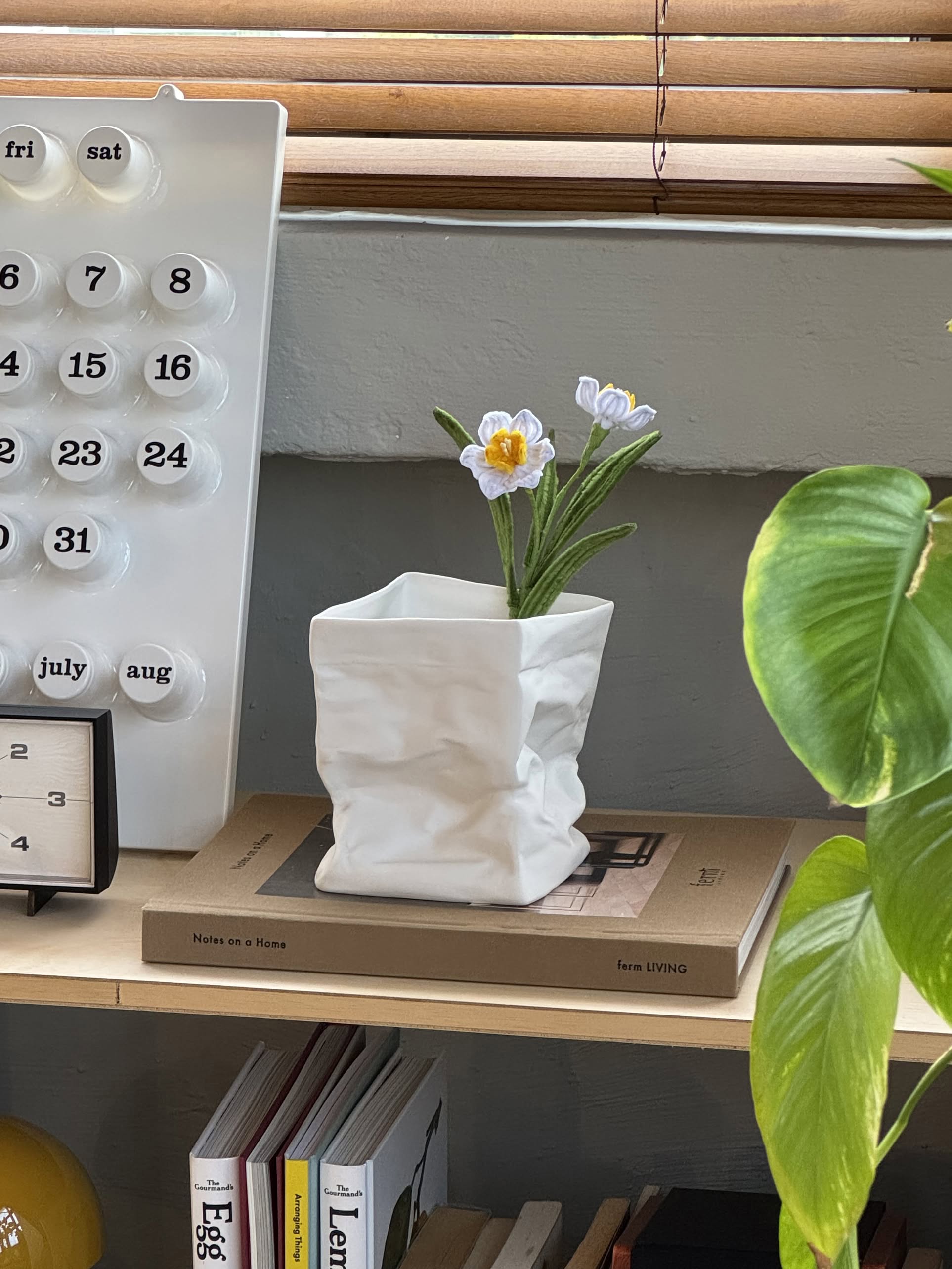 The Serax Kiki Paperbag L - White porcelain vase by Serax, with two artificial white flowers, sits on a book on a wooden shelf beside a green plant and under a wall calendar, bringing elegant Dutch design to the space.