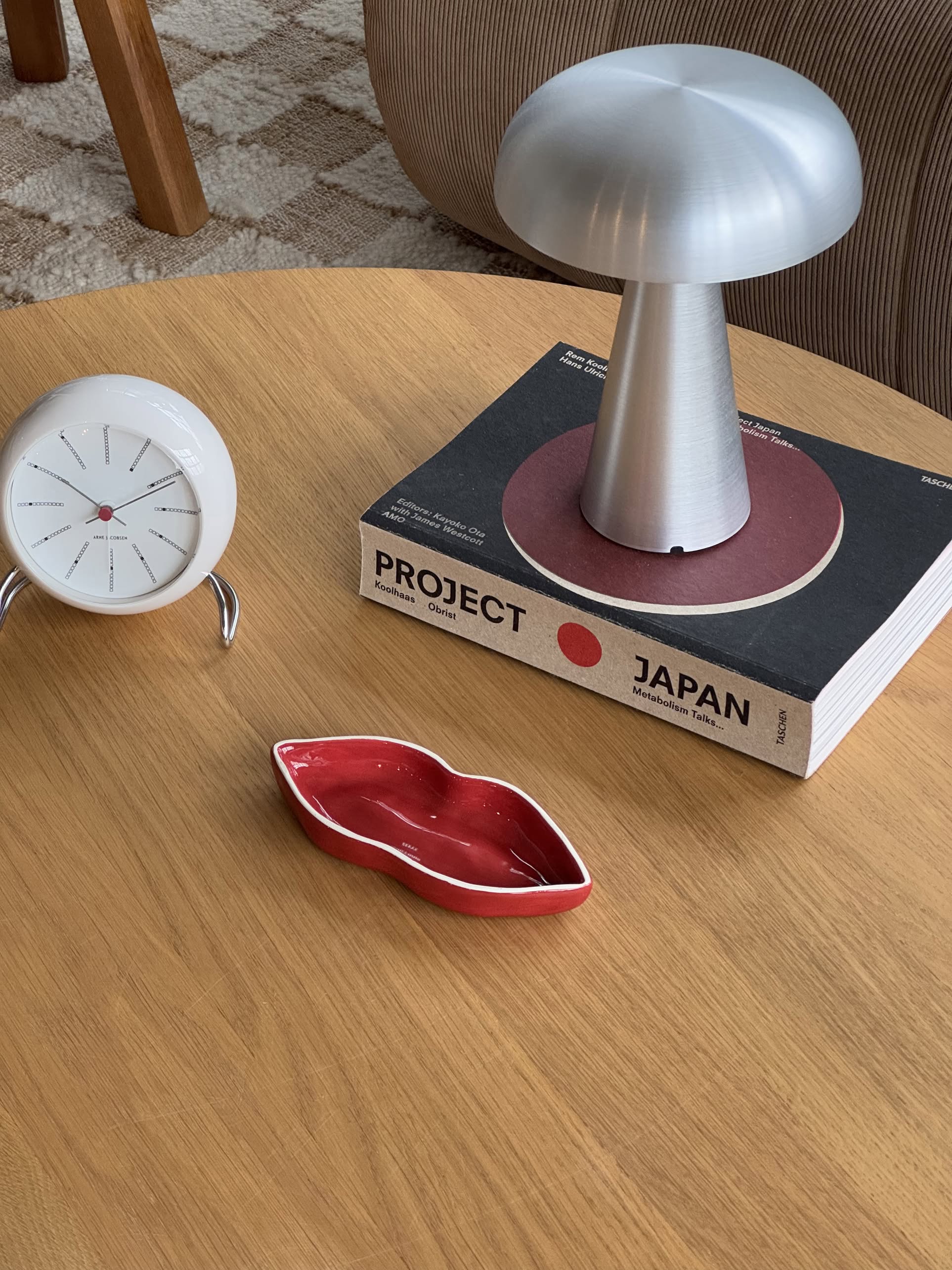 A wooden table features a white clock, the Serax Les Objets Mouleversants Serving Dish S - Red (lips-shaped) by Serax, a silver mushroom lamp, and the book PROJECT JAPAN with a red circle. A textured carpet and chair appear in the background.