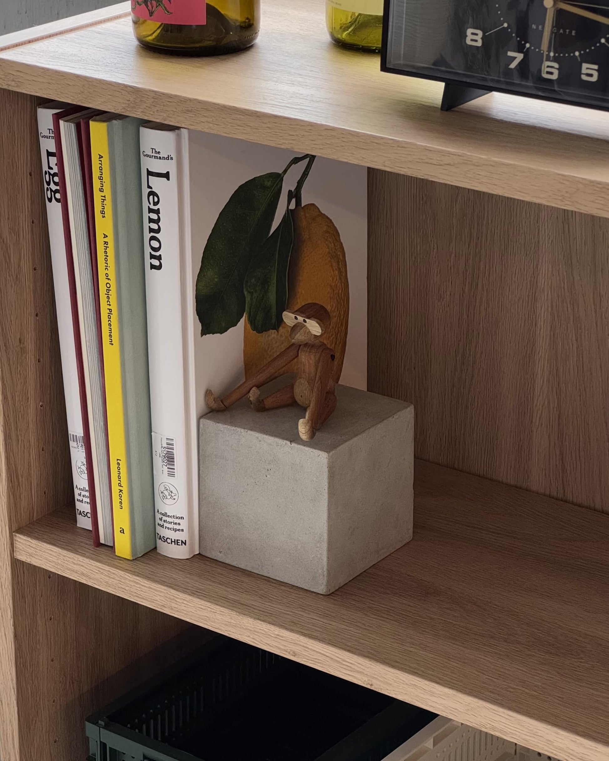 A wooden monkey figurine rests on a Serax Marie Cube Concrete - Grey by Serax, placed on a wooden shelf beside upright books—including one with a large lemon cover—while a clock and glass bottles decorate the space above.