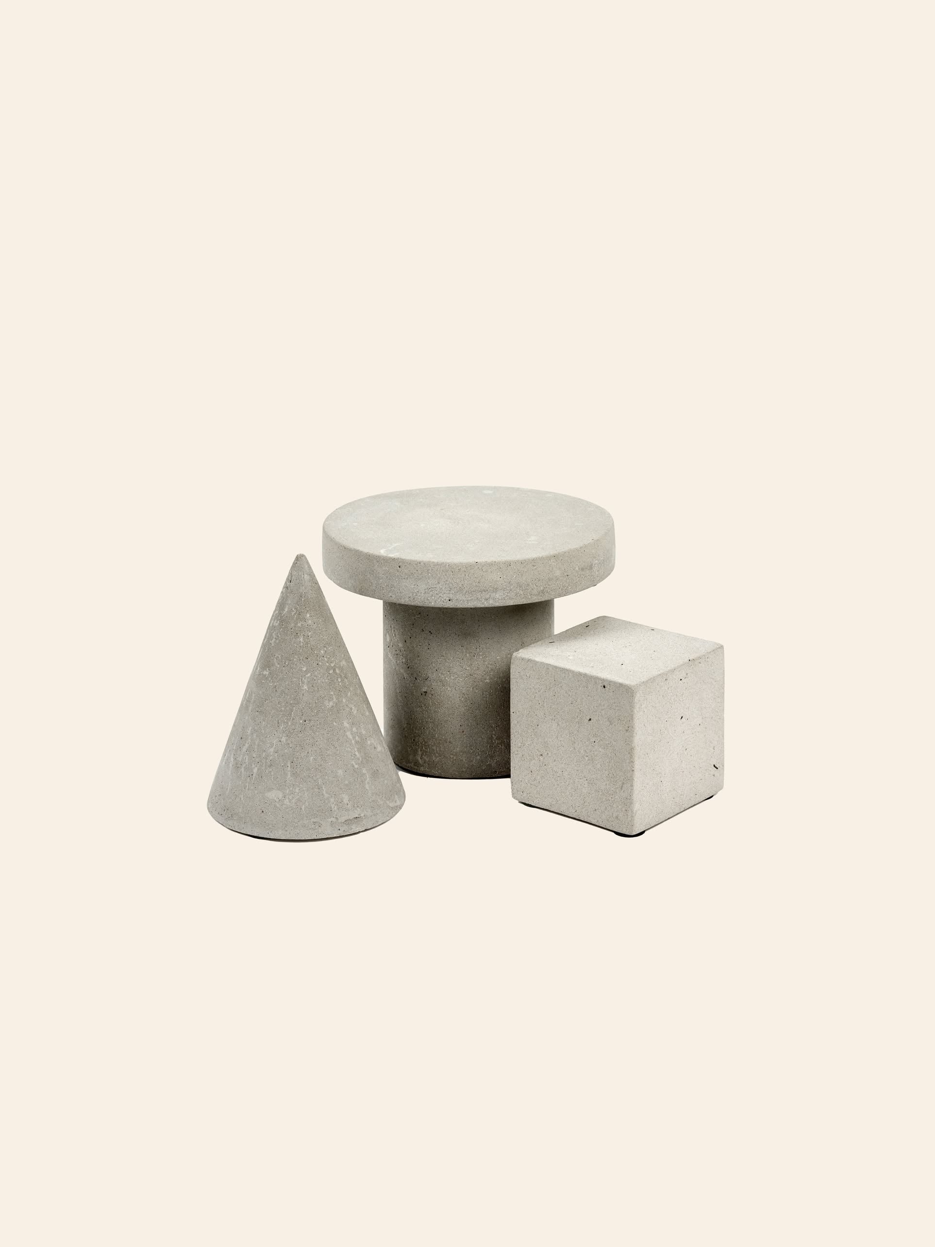 Three geometric shapes—a cone, a cylinder with a flat circular top, and the Serax Marie Cube Concrete - Grey by Serax—are arranged on a light beige background, lending an industrial vibe to modern decor.