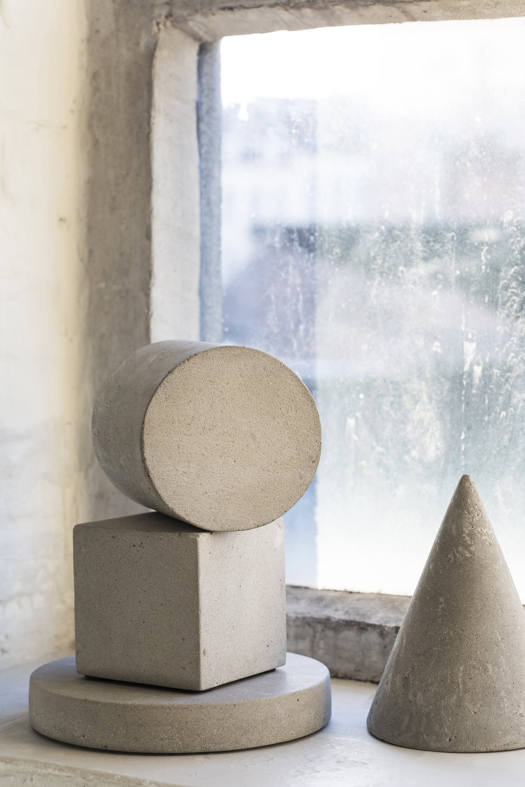 Concrete geometric shapes—a cylinder, a cone, a flat disk, and the Serax Marie Cube Concrete - Grey by Serax—are arranged on a windowsill beside a frosted window with natural light streaming in, lending an industrial touch to modern decor.