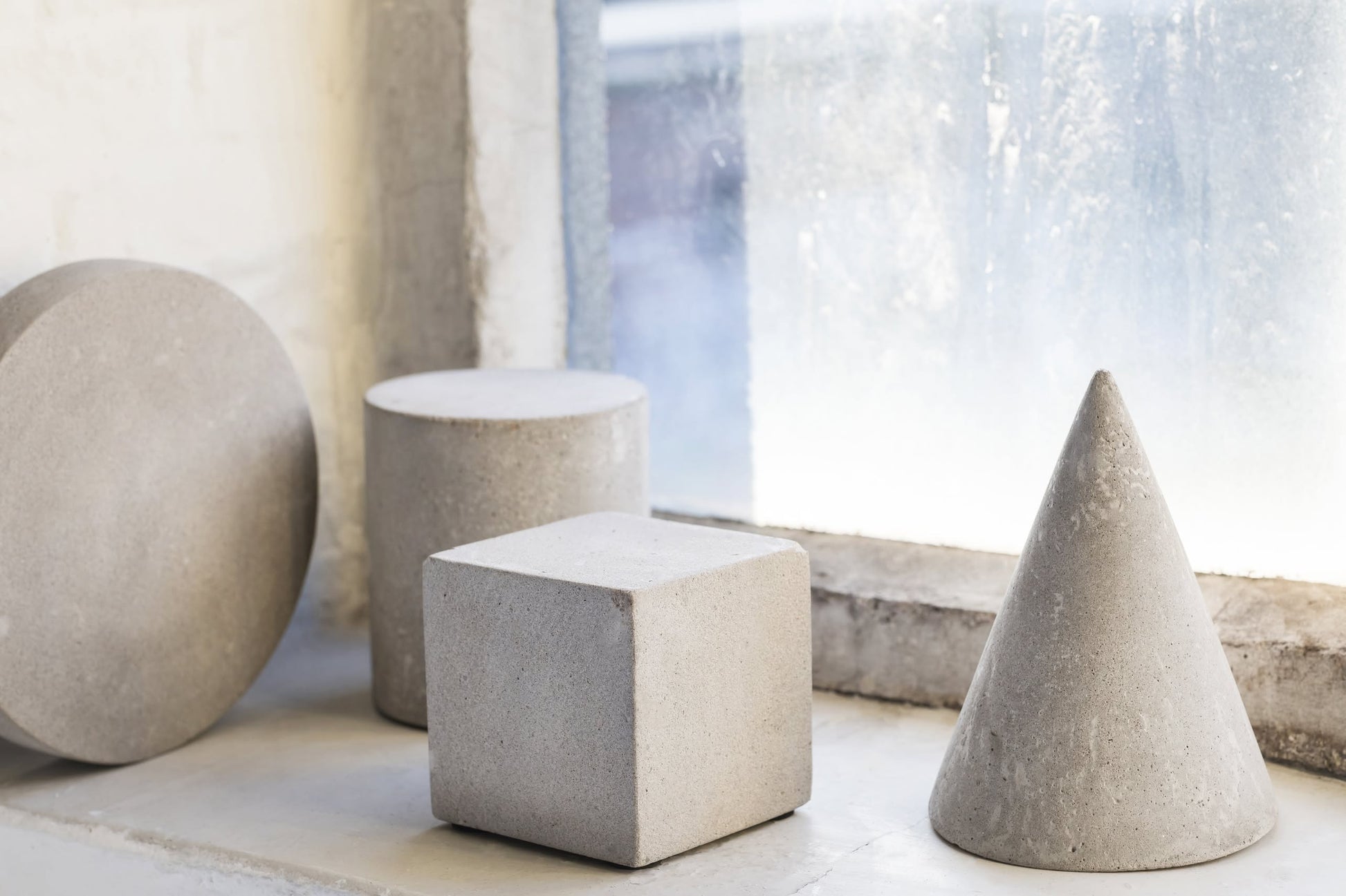 Four concrete geometric shapes—a cylinder, a sphere, the Serax Marie Cube Concrete - Grey, and a cone—sit on a windowsill before a frosty window, bringing an industrial accent to modern decor.