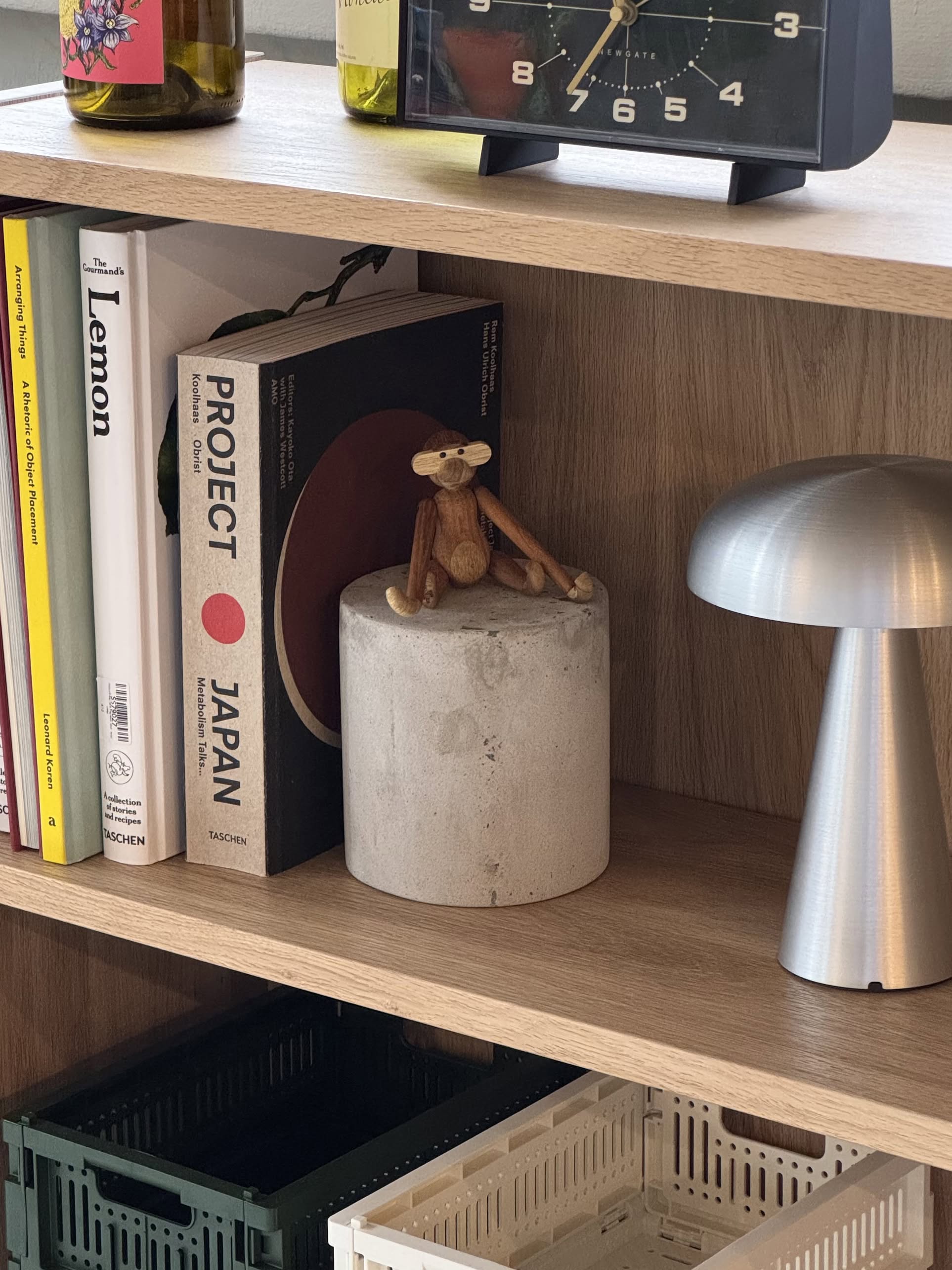 A wooden monkey figurine rests on the Serax Marie Cylinder Concrete - Grey by Serax, surrounded by books like PROJECT JAPAN and Lemon, plus a silver lamp—creating a minimalist yet industrial vibe on the wooden shelf.
