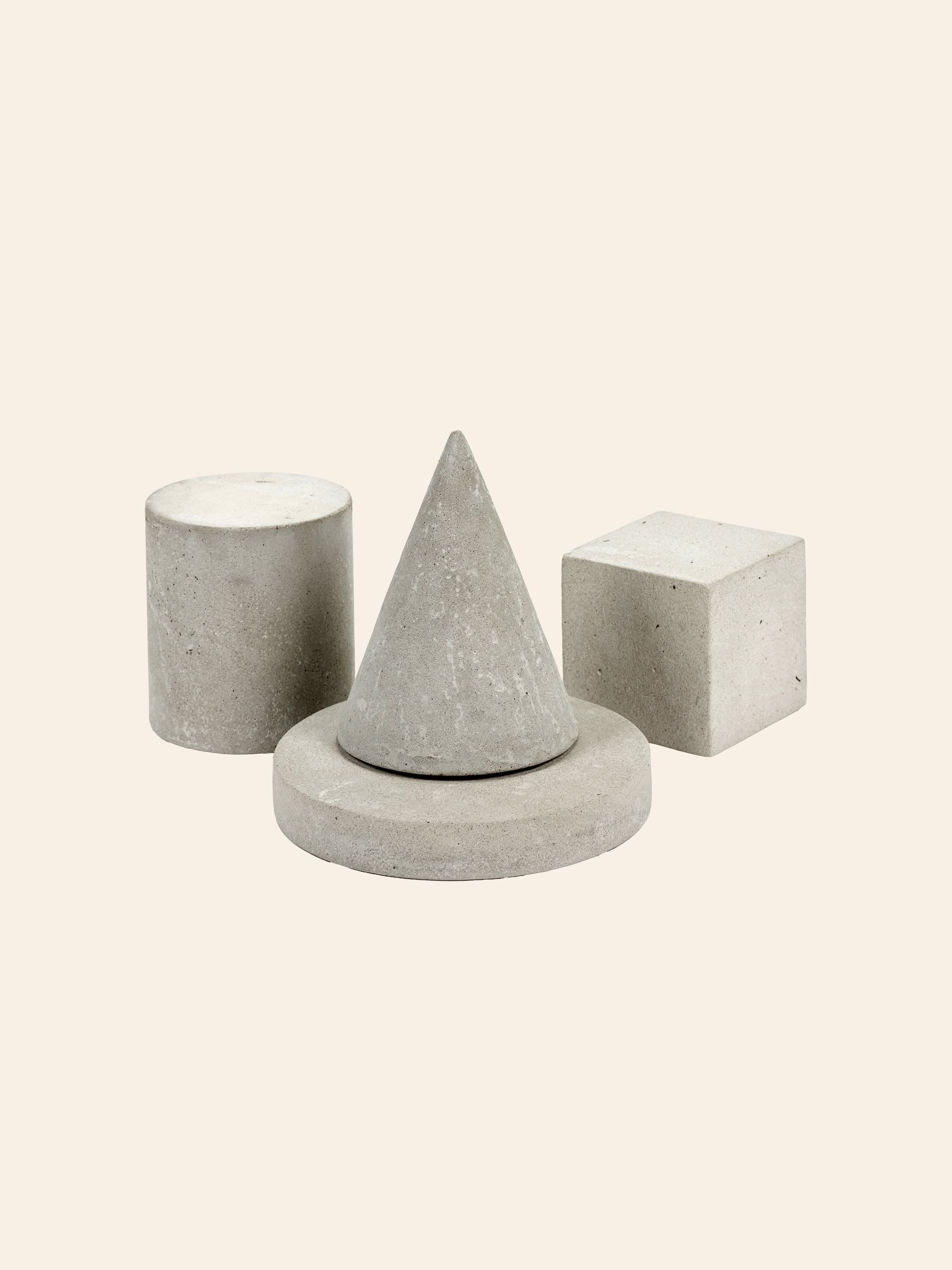 Four gray concrete geometric shapes—cylinder, cone, circular disc, and cube—are arranged on a light beige background. The Serax Marie Cylinder Concrete - Grey by Serax adds an industrial touch to the minimalist scene.