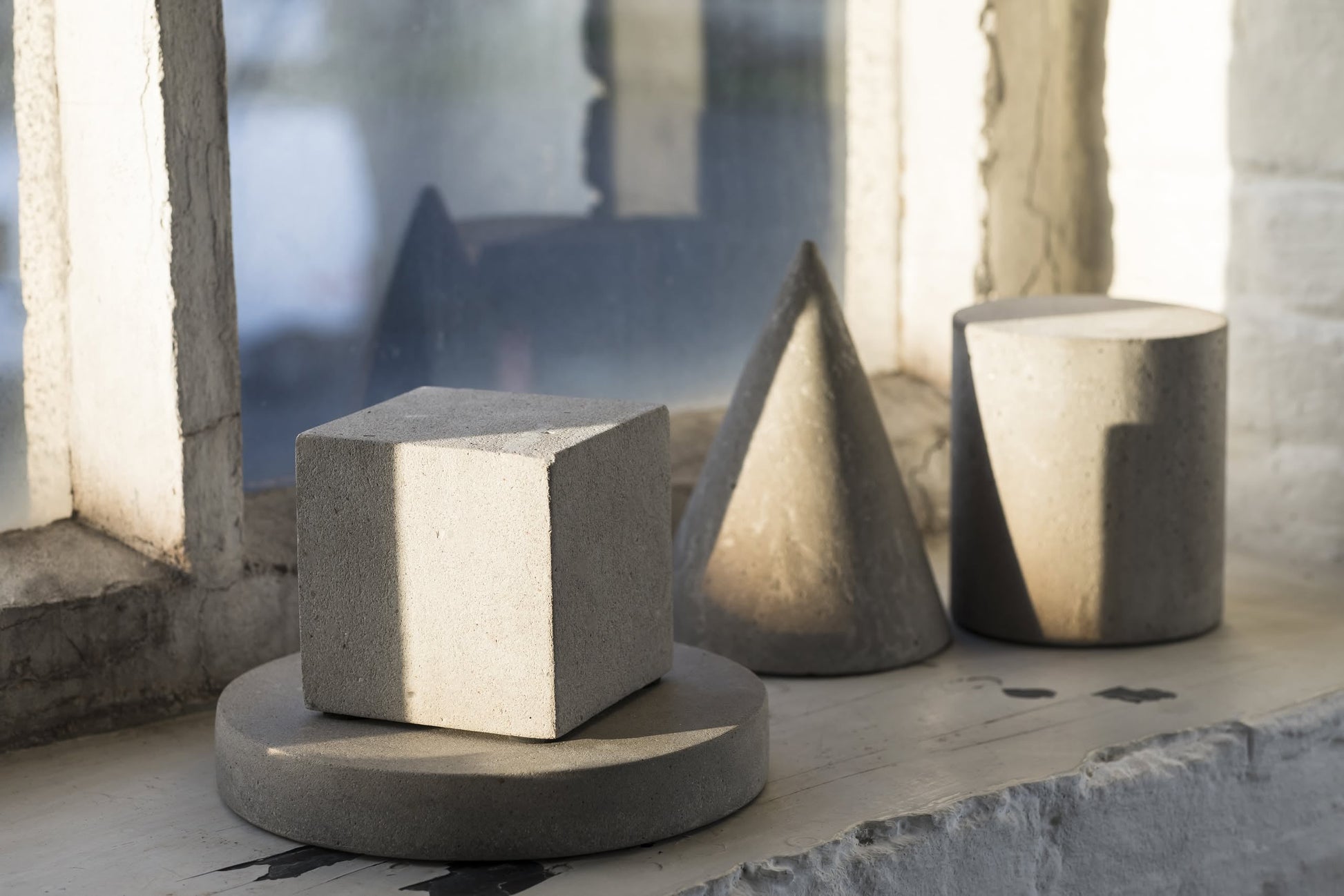 A cube, a cone, and the Serax Marie Cylinder Concrete - Grey by Serax rest on a windowsill in natural light, adding minimalist industrial style against the backdrop of an old, weathered window.