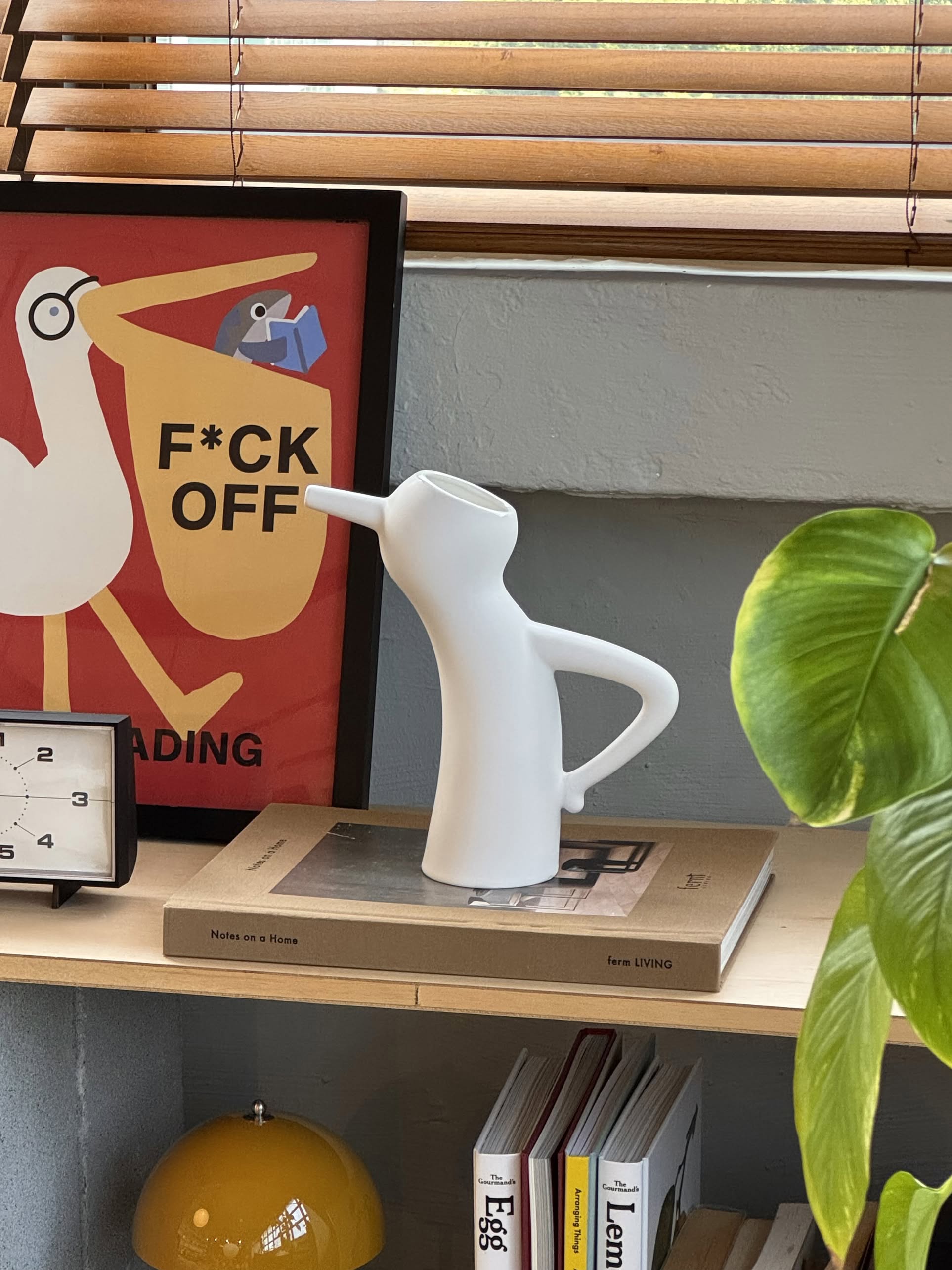 A Serax Monsieur Cruchot Jug S in white rests on a book atop a shelf. Behind it is a striking bird poster with “F*CK OFF” text, while a plant leaf appears in the foreground and a small clock sits to the shelf’s left.